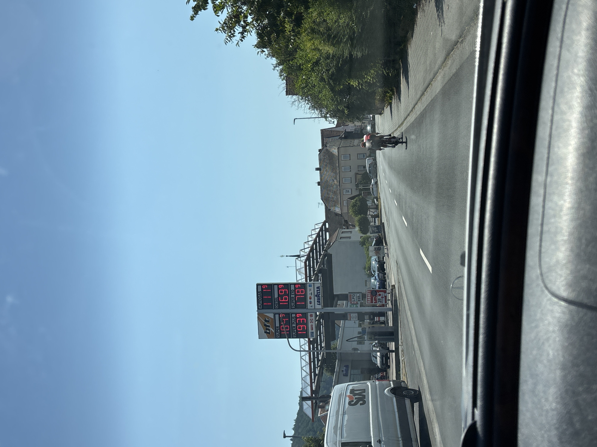 This is a street-level, eye-level, outdoor, daytime, candid photograph taken from the interior of a vehicle, looking forward through the windshield. The scene is a street in Wörth am Main, Germany. In the mid-ground to the left, a gas station with a prominent price display is visible. The price display shows current fuel prices. The prices listed are: AUTOGAS 1.119, DIESEL 1.699, SUPER 1.849, SUPER E10 1.789, and SUPER PLUS 1.939. To the left of the gas station, a white van with "Sixt" branding is parked. Cars are lined up at the gas station. Two cyclists are riding on the right side of the road in the mid-ground, one in a red shirt and the other in a darker shirt. The road is paved and has lane markings. Buildings, trees, and a blue sky with a few clouds are visible in the background. The foreground shows the interior of the vehicle's dashboard.