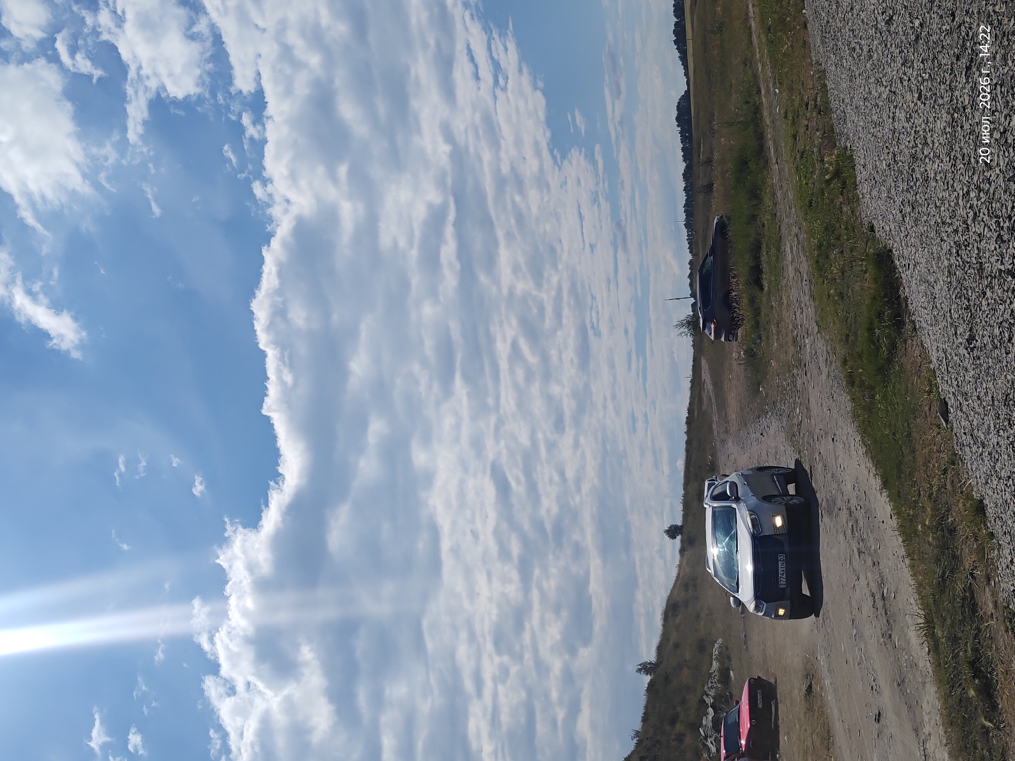 In the city of Akylbay, Kazakhstan, on July 20, 2026, at 14:22, a scene unfolds on a dirt road under a partly cloudy sky with bright sunlight filtering through the clouds. Three cars are visible: a red car is parked on the left side of the road, a silver SUV is driving towards the viewer on the road, and a dark-colored SUV is parked further ahead on the right side of the road, off the main path. The terrain is dry and dusty, with sparse vegetation on the surrounding hills. The ground is covered with gravel in the foreground, indicating a possible construction or roadside area. The overall impression is a rural or semi-rural environment, possibly a recreational area or a less-developed road.