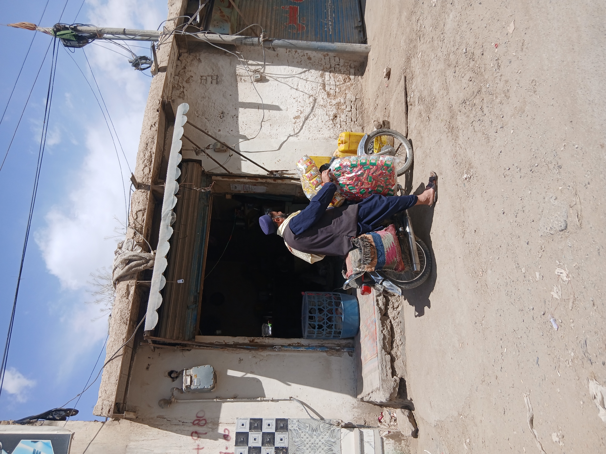 A man is riding a bicycle loaded with goods in Quetta City Tehsil, Pakistan. He is wearing a dark blue coat and a light-colored head covering. The bicycle has a large amount of packaged food and other items loaded onto it. The man is in front of a building with a weathered awning. The building shows signs of wear and age. The ground is dusty. There is a utility pole and wires visible in the upper left of the image.