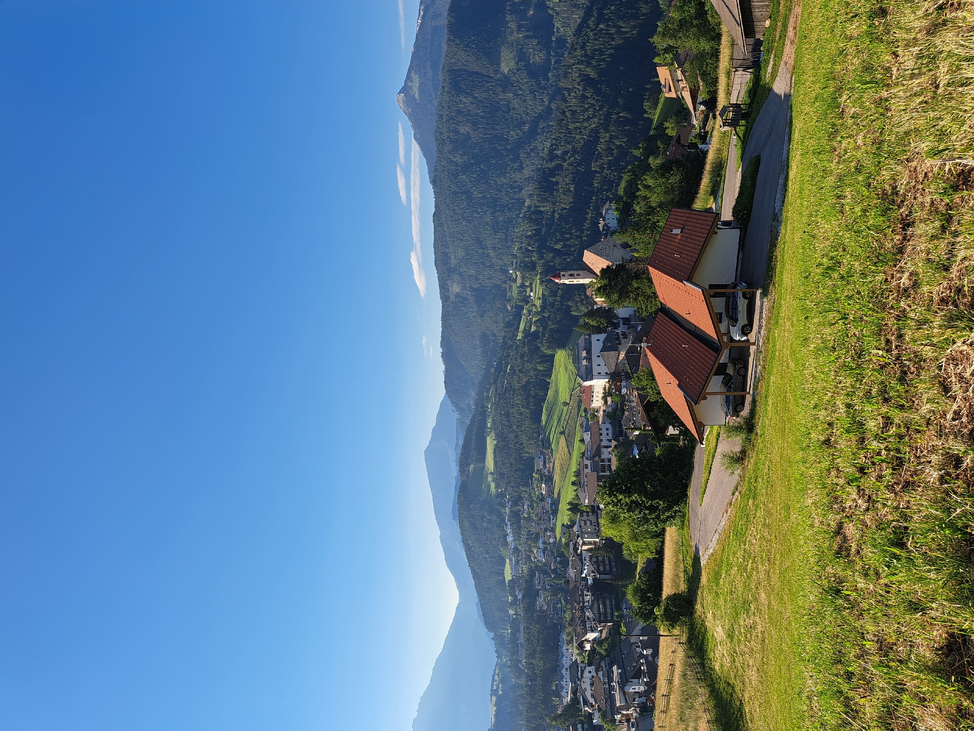 The image captures a panoramic view of a mountainous village bathed in the warm light of a clear day. A vast expanse of blue sky dominates the left side of the frame, gradually giving way to layers of distant, misty mountains. The closer mountains are covered in dense, dark green forests, creating a textured backdrop for the village nestled in the valley.
The village itself is a cluster of buildings with red-tiled roofs, interspersed with patches of green fields and trees. Several houses and structures are visible, some with white or light-colored walls. One house in the foreground has a covered parking area with two cars parked underneath. A paved road winds its way through the village and up the hillside.
The foreground is dominated by a grassy slope, with the grass appearing somewhat dry and golden in the sunlight. The overall scene suggests a peaceful, rural setting in a mountainous region, likely during the daytime given the bright sunlight and clear skies. The light creates long shadows, indicating it might be late afternoon or early morning. There is no visible text in the image.