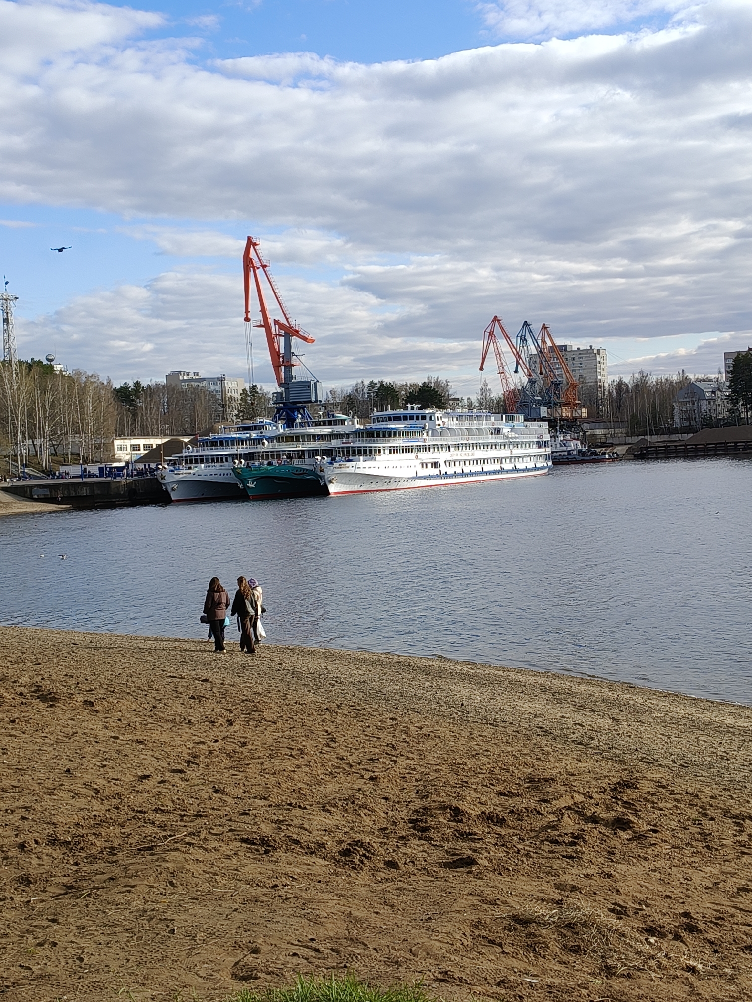 The image depicts a waterfront scene, most likely in the city of Chaikovsky, Russia, given the architectural style and presence of river cruise ships. In the foreground, a sandy beach is visible, with three people standing near the water's edge. The middle ground shows a body of water with several large river cruise ships docked at a pier. In the background, cranes rise above the ships, suggesting a port or industrial area. Buildings and trees line the opposite bank, partially obscuring the urban landscape. The sky is filled with clouds, indicating a slightly overcast day. A bird is flying in the upper left corner of the image.
