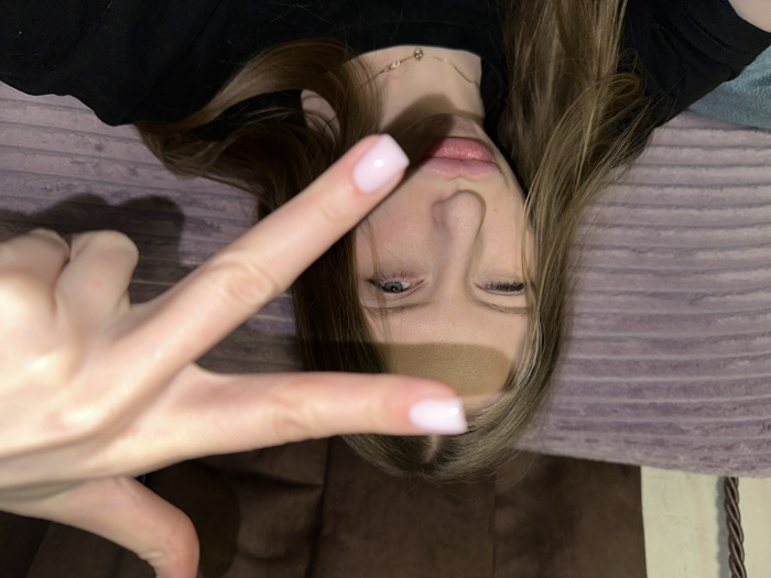 The image displays a young female subject indoors, located within Chaikovsky, Russia; the specific address or building is not discernible. The subject's face, positioned centrally, exhibits a winking or squinting left eye and pouted lips. A light-skinned hand, presumably belonging to the subject, is extended into the foreground, with the index finger horizontally across her forehead and the thumb vertically along her right cheek, partially obscuring her face. The fingernails visible on the hand feature light pink nail polish. A shadow cast by the hand is prominent across the subject's forehead and nose. She is wearing a black top and a thin, gold-colored chain necklace with a small pendant. The background consists of soft furnishings: a light purple, horizontally textured fabric occupies the left side, and a dark brown, plush-like fabric is on the right, indicative of a domestic setting.