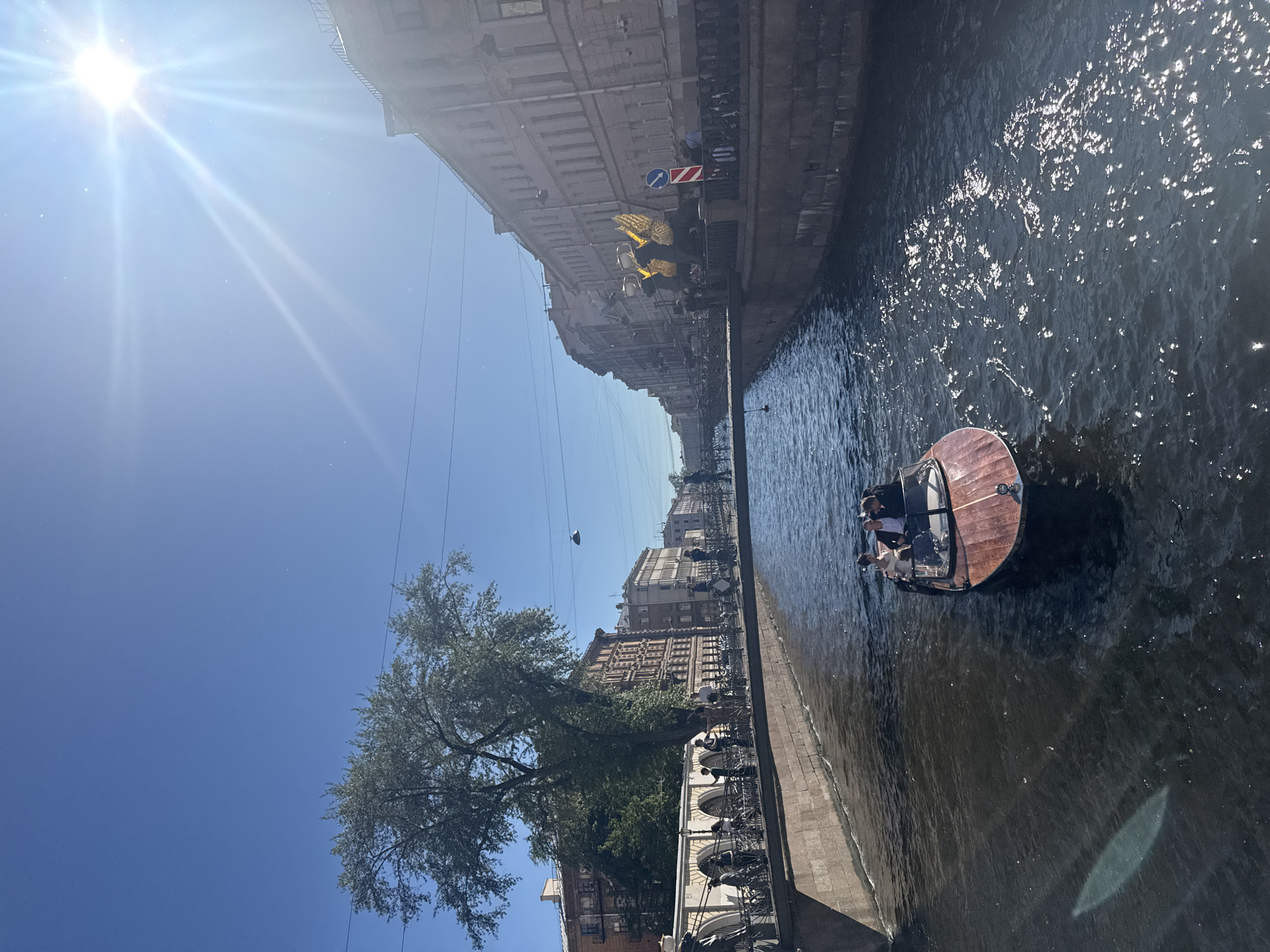 The image depicts a sunny day in Saint Petersburg, Russia, likely along one of its many canals. In the foreground, a wooden boat with a modern design navigates the dark, rippling water. Several people are seated in the boat, appearing to be on a tour or enjoying a leisurely ride. The canal is flanked by stone embankments, with pedestrian walkways running alongside. A bridge spans the canal in the mid-ground, populated with people. On the right bank, tall, multi-story buildings with classical architecture line the street. The sky is a bright, clear blue with a prominent sun, casting lens flares and reflections on the water. On the left bank, a large tree with lush green foliage stands tall, with more buildings visible behind it. Further down the canal, more bridges and buildings are visible, suggesting an urban setting with a rich architectural history. The overall scene conveys a vibrant and picturesque atmosphere.