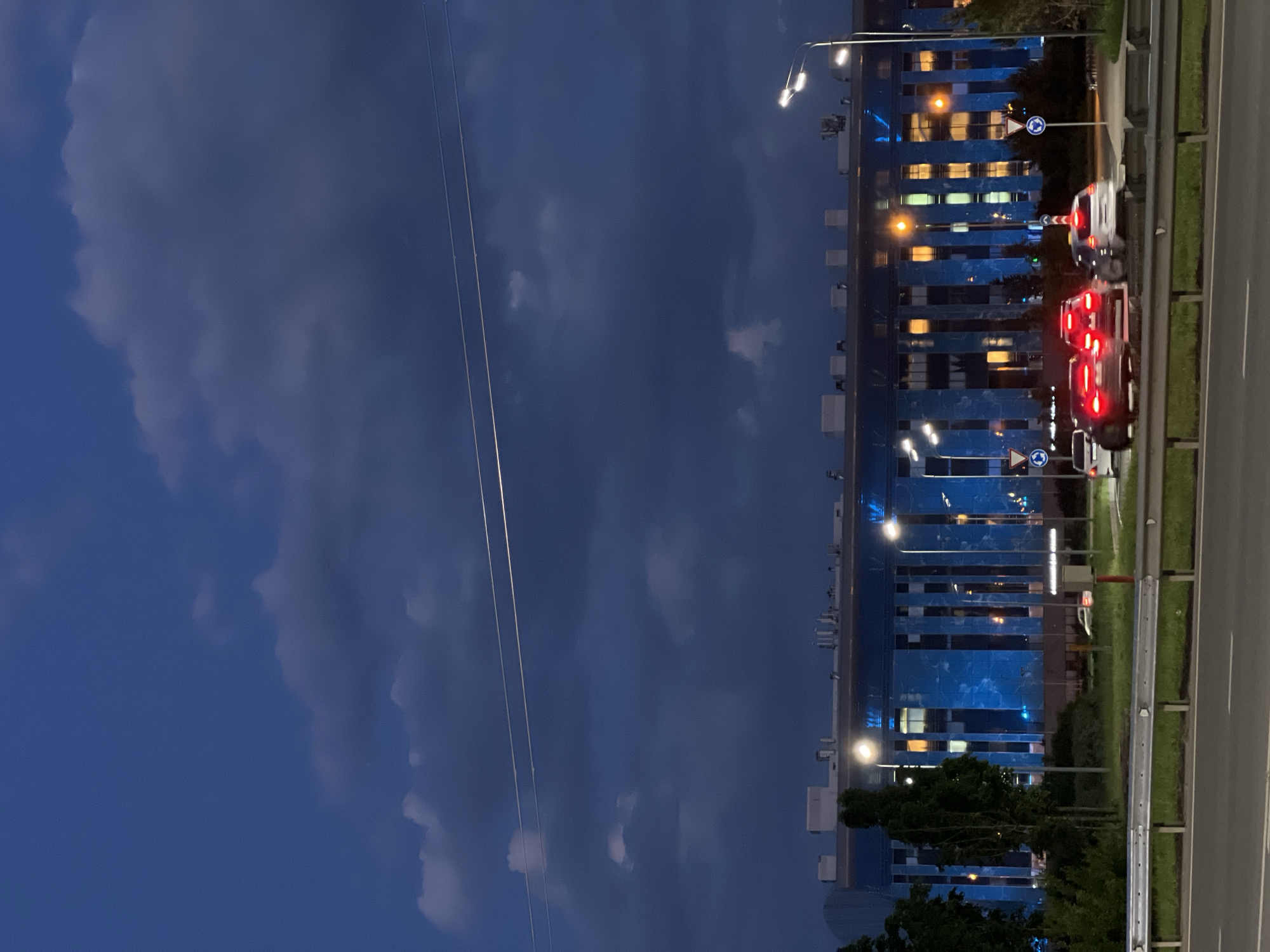 An exterior shot of a blue-paneled building in Dachnoe, Russia, is captured at dusk. The building features numerous windows, some of which are illuminated with warm-toned light, contrasting with the cool blue exterior. Several streetlights are visible, casting a glow on the scene. A highway in the foreground shows a few cars in motion, their headlights and taillights creating streaks of light due to the long exposure. The sky above is a deep blue, with a few scattered clouds. Trees and greenery line the area between the highway and the building.