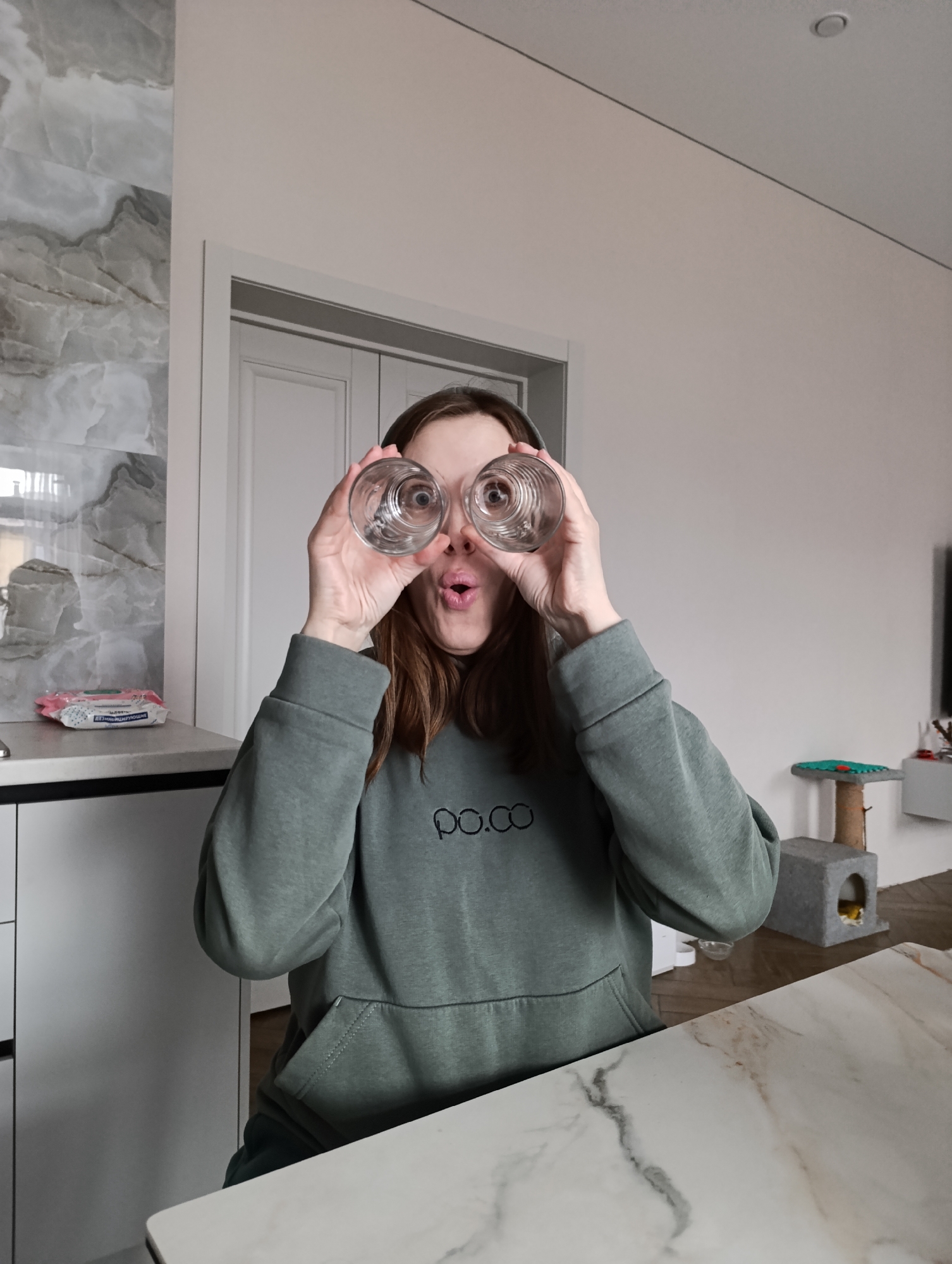 A woman is sitting at a marble countertop in what appears to be a kitchen. She is holding two empty, clear drinking glasses in front of her eyes, as if they were binoculars, and making a surprised or playful facial expression. She is wearing a green hooded sweatshirt with the word "po.co" embroidered on the front. Behind her are white cabinets, a white doorway, and part of a cat tree. A package of wet wipes is visible on the countertop to her left. The precise location in the city of Chaikovsky, Russia, cannot be determined from the image.