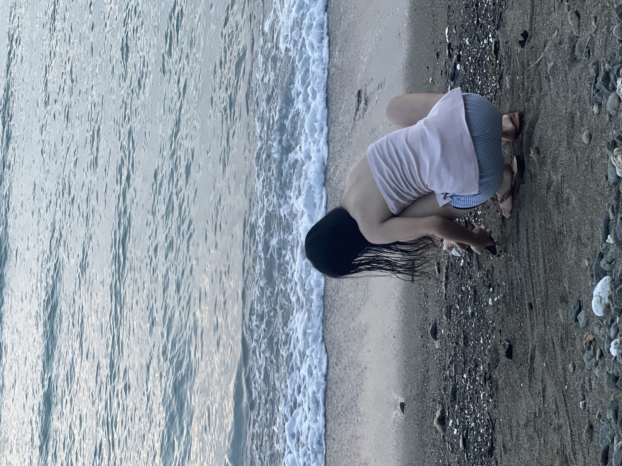 A person with long, dark hair is bent over, collecting pebbles on a beach in Banaybanay, Philippines. They are wearing a light pink shirt and striped shorts and are standing near the edge of the water where the waves are foaming. The sand on the beach transitions from light to dark, with pebbles scattered across the darker section. The sea occupies the left side of the image.