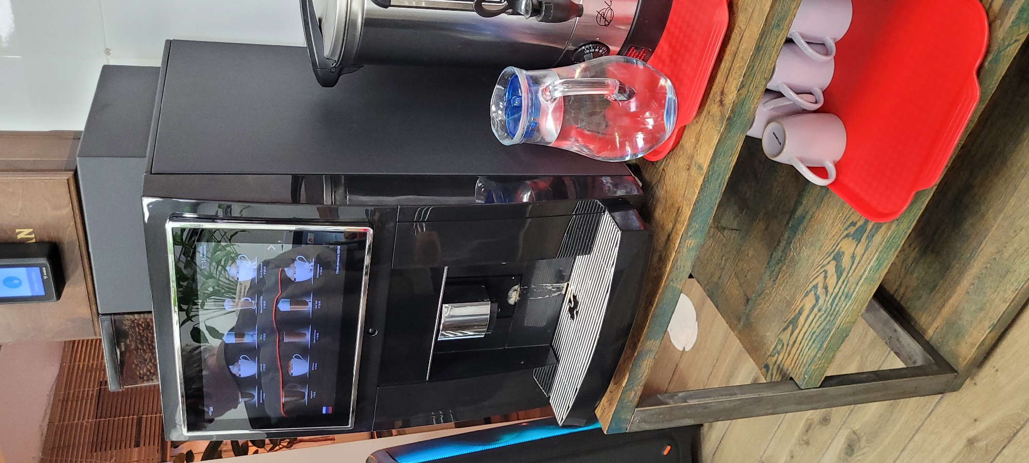 Here is a detailed description of the image:
The scene features a black automated coffee machine resting on a wooden surface, likely a countertop or shelf. A clear glass pitcher filled with water sits to the right of the coffee machine, adjacent to a red tray holding several white coffee cups. The coffee machine has a digital display showing different coffee selections. A small electronic device is visible to the left of the coffee machine on a lower surface. The wooden surface appears to be part of a structure with metal framing.