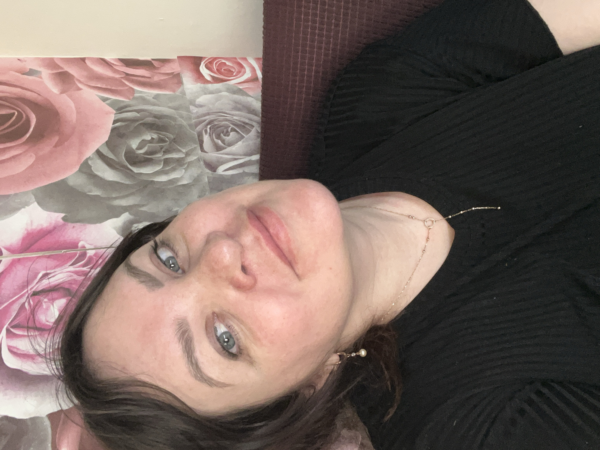 A woman lies on a floral patterned surface next to a textured, maroon-colored surface. The woman is wearing a black, ribbed top and a gold necklace with a pendant, and is looking upwards with blue eyes. She has brown hair and appears to be indoors. The background is decorated with a pink and gray rose pattern. The location is not verifiable as Noviy, Russia based on the provided image.