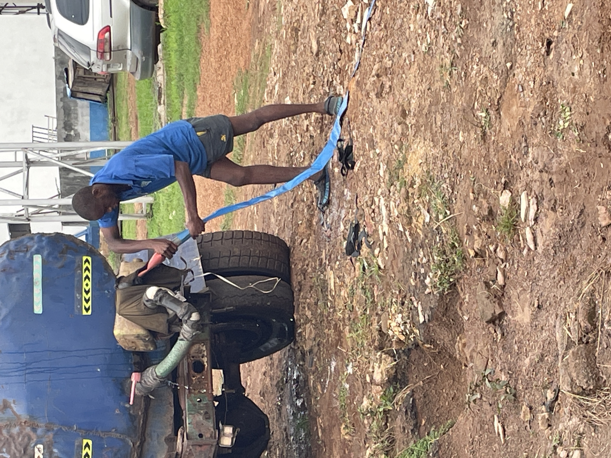 In Enugu, Nigeria, a man in a blue t-shirt and gray shorts is seen holding a blue hose connected to a large, weathered blue water tank truck. The hose appears to be dispensing water onto the muddy, uneven ground, which is littered with small rocks and sparse vegetation. The truck is positioned on a dirt area, with its tires visible. In the background, a silver SUV is parked, and a metal structure, possibly part of a building or industrial equipment, stands against a pale wall. The overall environment suggests an outdoor, possibly industrial or service area.