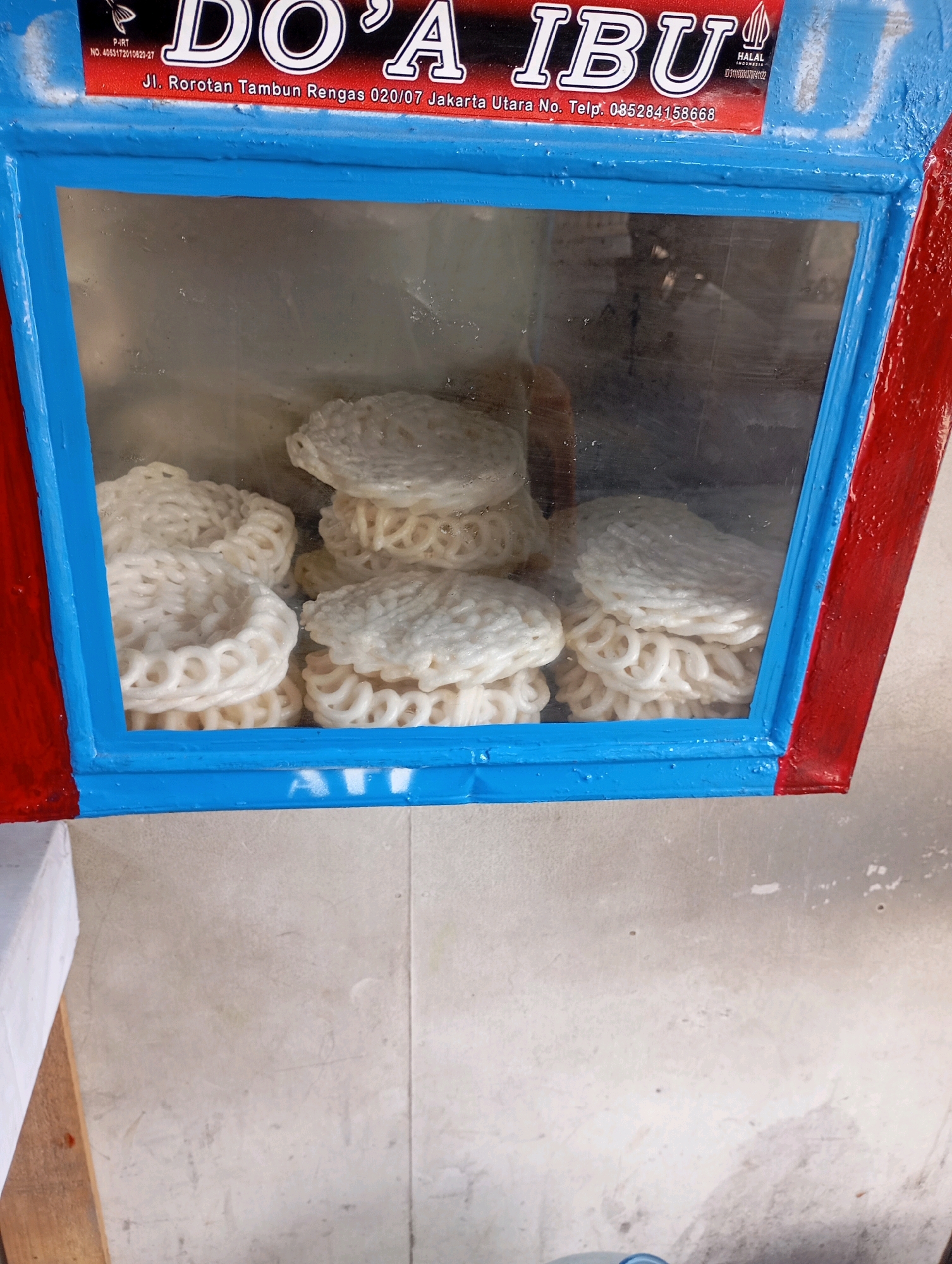 A close-up, eye-level shot shows a display case containing stacks of white, ring-shaped crackers. The display case has a blue frame and a red border. Above the display case, a sign reads "DO'A IBU" in large white letters with a black outline. Below the title, text in smaller white letters indicates the location as "Jl. Rorotan Tambun Rengas 020/07 Jakarta Utara No. Telp. 085284158668". To the right of "DO'A IBU" is a "HALAL" logo. The background appears to be a wall with a rough texture.
