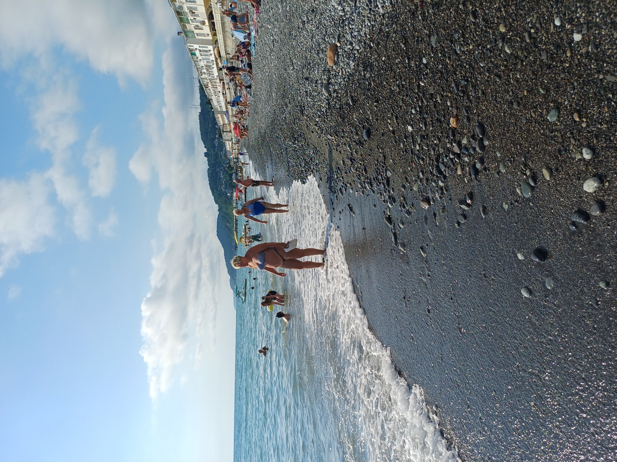 The image depicts a crowded beach on a sunny day in Sochi, Russia. The beach is covered in dark pebbles, and the sea is calm with gentle waves lapping at the shore. Several people are visible in the water, including adults and children swimming and playing. On the beach, individuals are sunbathing, sitting on towels, and walking along the shoreline. In the background, a row of white buildings with balconies lines the coast, suggesting a resort area. The sky is blue with scattered white clouds, and a lush green hillside rises behind the buildings, characteristic of Sochi's coastal landscape. The overall scene conveys a typical summer day at a popular tourist destination.