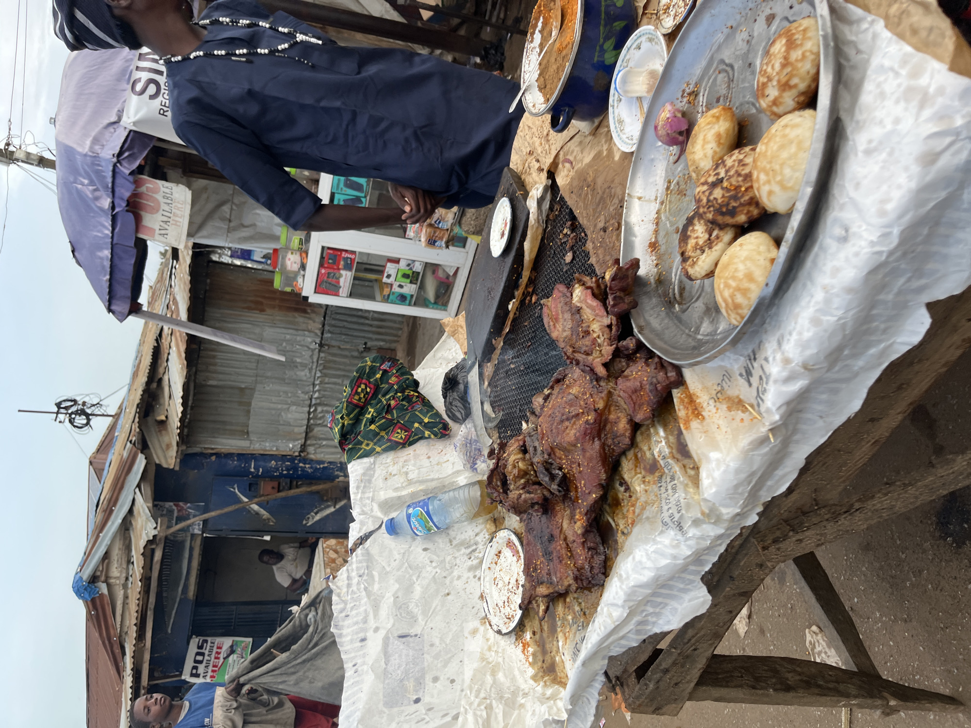 The image depicts an outdoor food stall in Bukuru, Nigeria. In the foreground, a wooden table displays a spread of food. Large cuts of grilled meat, seasoned with spices, are arranged on a white paper liner. To the right of the meat is a silver platter containing several round, browned pastries, possibly pancakes or a similar local delicacy. A blue pot containing a reddish-brown stew or sauce sits on the table, with a spoon resting inside. Smaller plates with remnants of food and a red onion are also visible.
In the background, a young boy in a blue shirt holds a grey cloth. Behind him, a stall with a corrugated metal roof and blue walls is visible. A sign in English reading "POS AVAILABLE HERE" is prominently displayed. Further back, a man in a dark blue traditional outfit with beaded necklaces is seen standing. The environment suggests a bustling market or street setting with various stalls and structures.