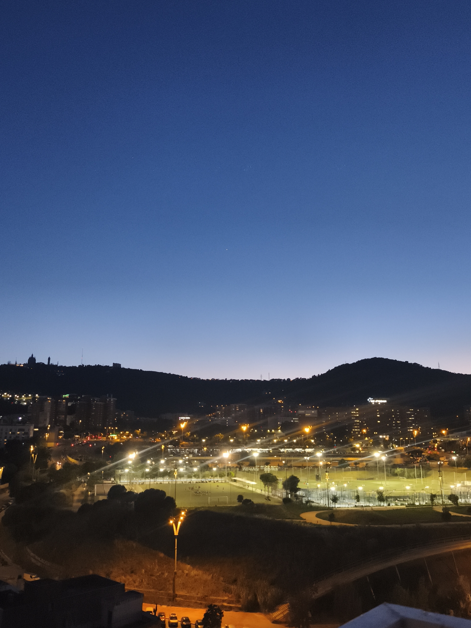 A nighttime shot captures a city scene, likely Barcelona, Spain, given the architectural style and hilly backdrop. The dark blue sky transitions to a lighter horizon near the silhouette of distant hills. The city below is lit with warm streetlights, illuminating buildings, streets, and what appears to be a park or recreational area with lit sports fields. There are no people visible.