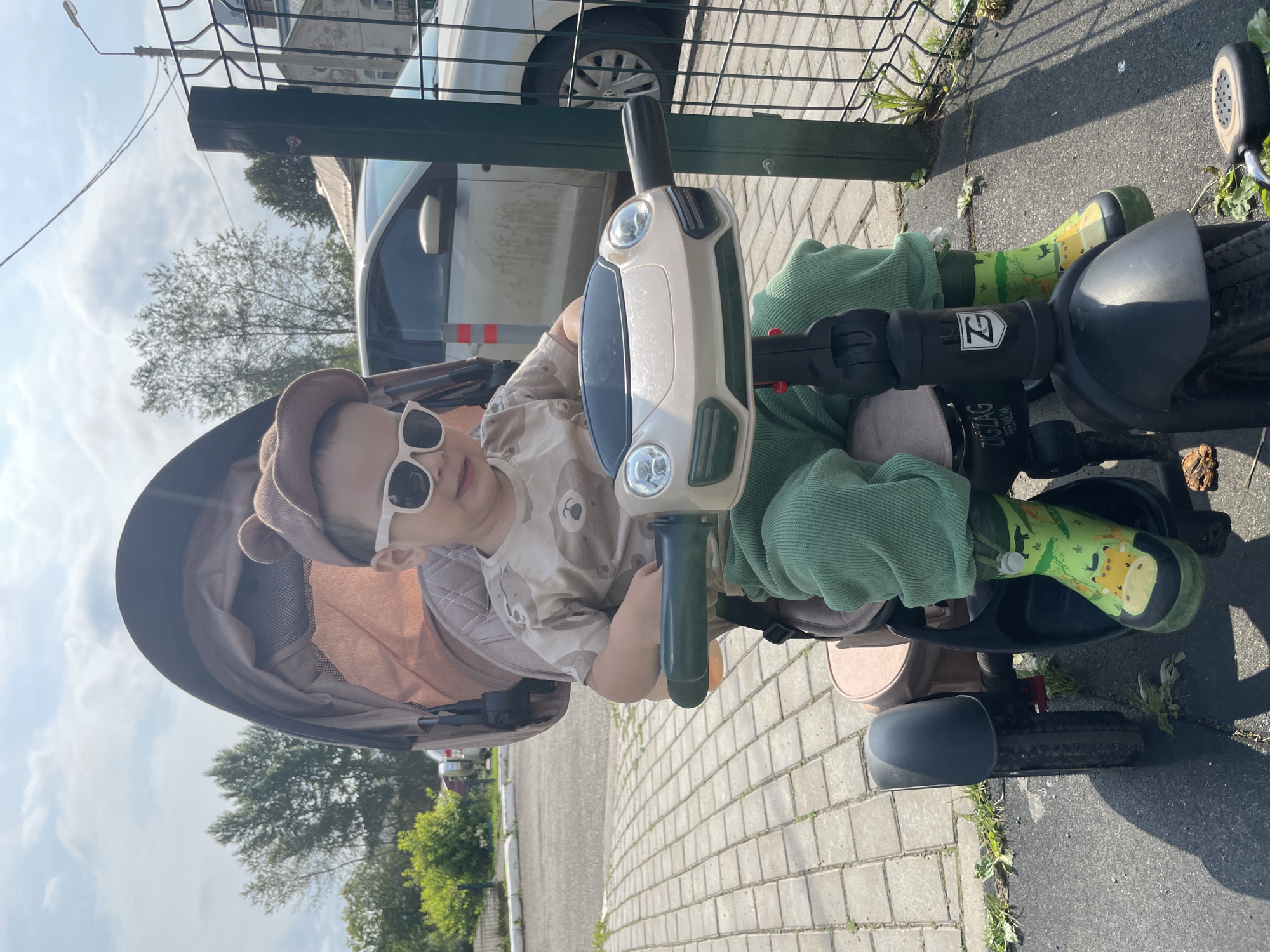 A young child, wearing a bear-ear hat, sunglasses, a light brown t-shirt with a bear design, green pants, and green rain boots with animal prints, sits on a tricycle. The tricycle has a light beige and black body, with a green handlebar. Behind the child is a stroller with a beige canopy. The child is facing forward, towards the viewer. The tricycle is parked on a paved area made of grey interlocking bricks, with shadows cast on the ground. In the background, there is a green metal fence, a light-colored car, some trees, and buildings. The sky is blue with some clouds, indicating an outdoor setting during the day.