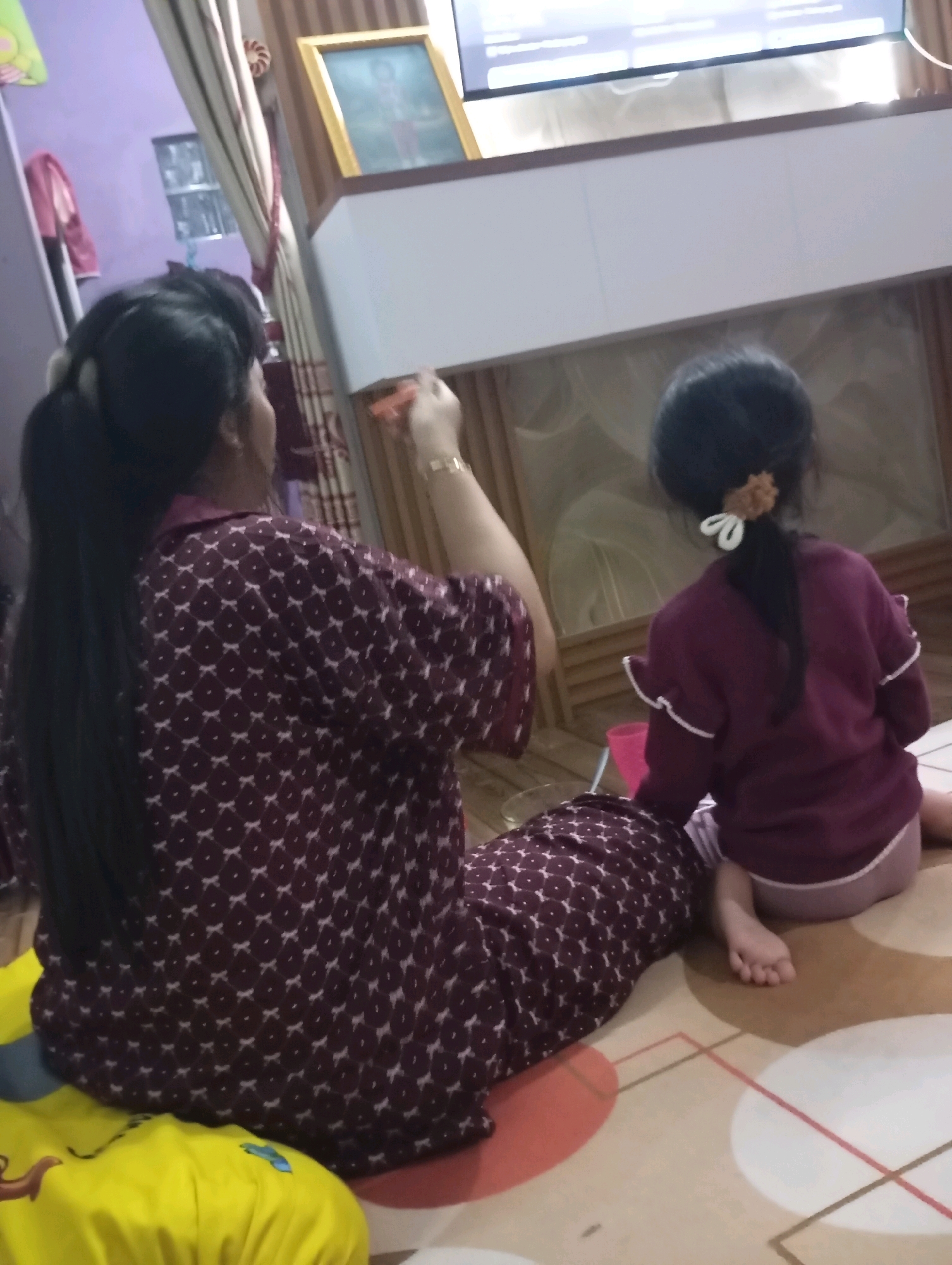 The image depicts a domestic scene in Muncang, Indonesia. Two individuals, an adult and a child, are seated on a patterned carpeted floor. The adult, facing away from the camera, has long dark hair tied into a ponytail and is wearing a dark maroon pajama set with a white geometric pattern. They appear to be holding a small orange object in their right hand, which is adorned with a bracelet. The child, also facing away from the camera, has dark hair styled in a ponytail with a light brown scrunchie and a white ribbon. They are wearing a dark maroon long-sleeved shirt with white trim and light purple shorts. The child's legs are visible, with their bare feet resting on the carpet.
In the background, a television screen displays content, suggesting the individuals are watching something. To the left, a framed picture is visible on a wooden structure, and curtains hang in the background. The environment appears to be a living room or a similar indoor space within a residence. The overall lighting is dim, with some light emanating from the television. The carpet has a circular pattern with red and white elements. A pink cup is partially visible near the child.