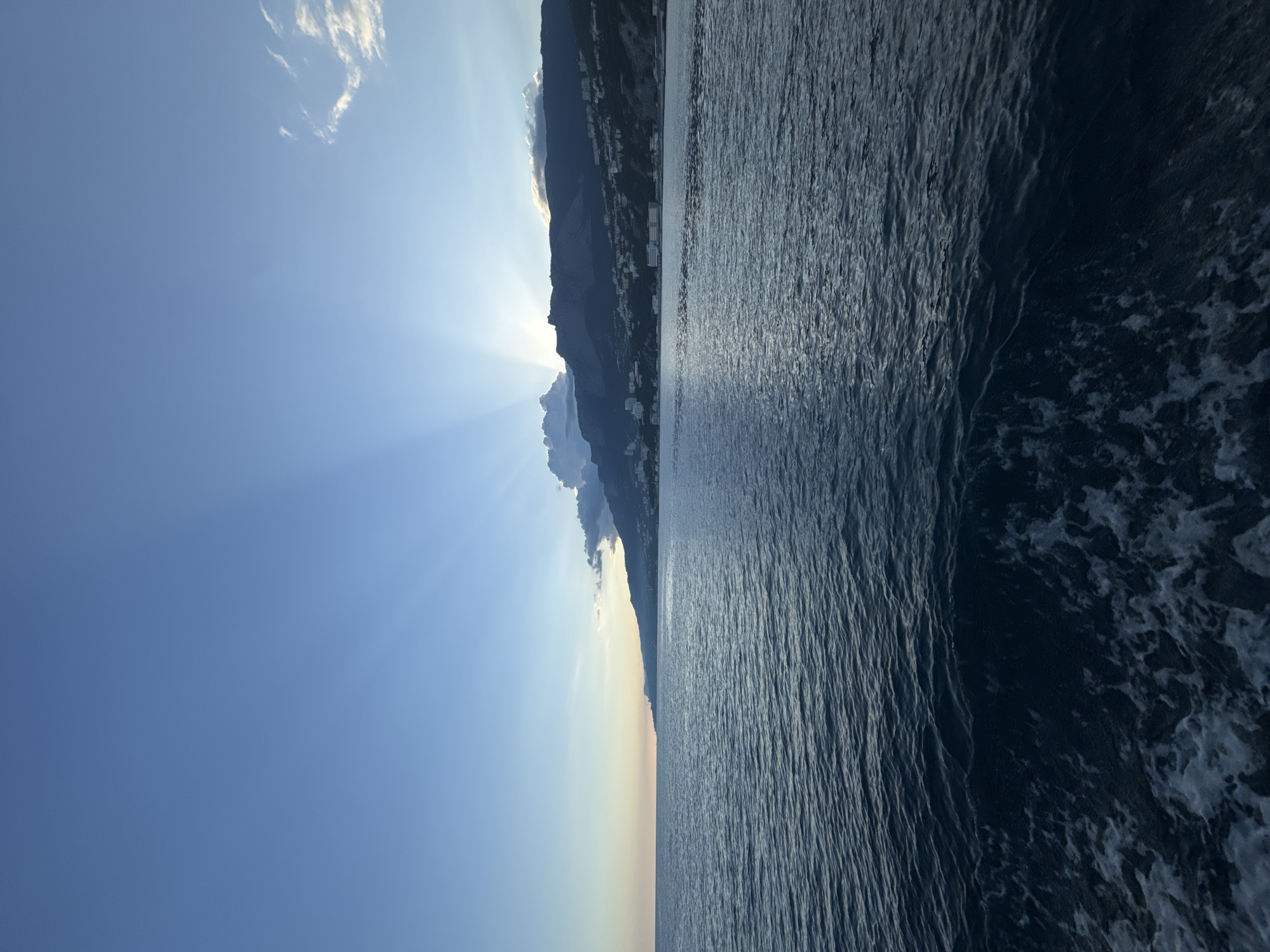 The image shows a view of the sea and a mountain range in Гаспринский поселковый совет, Ukraine. The sea is on the right, with dark blue water and some white foam visible. The mountain range is on the left, with several buildings visible on the slopes. The sky is blue, with some clouds, and the sun is shining behind the mountains, creating a ray effect.