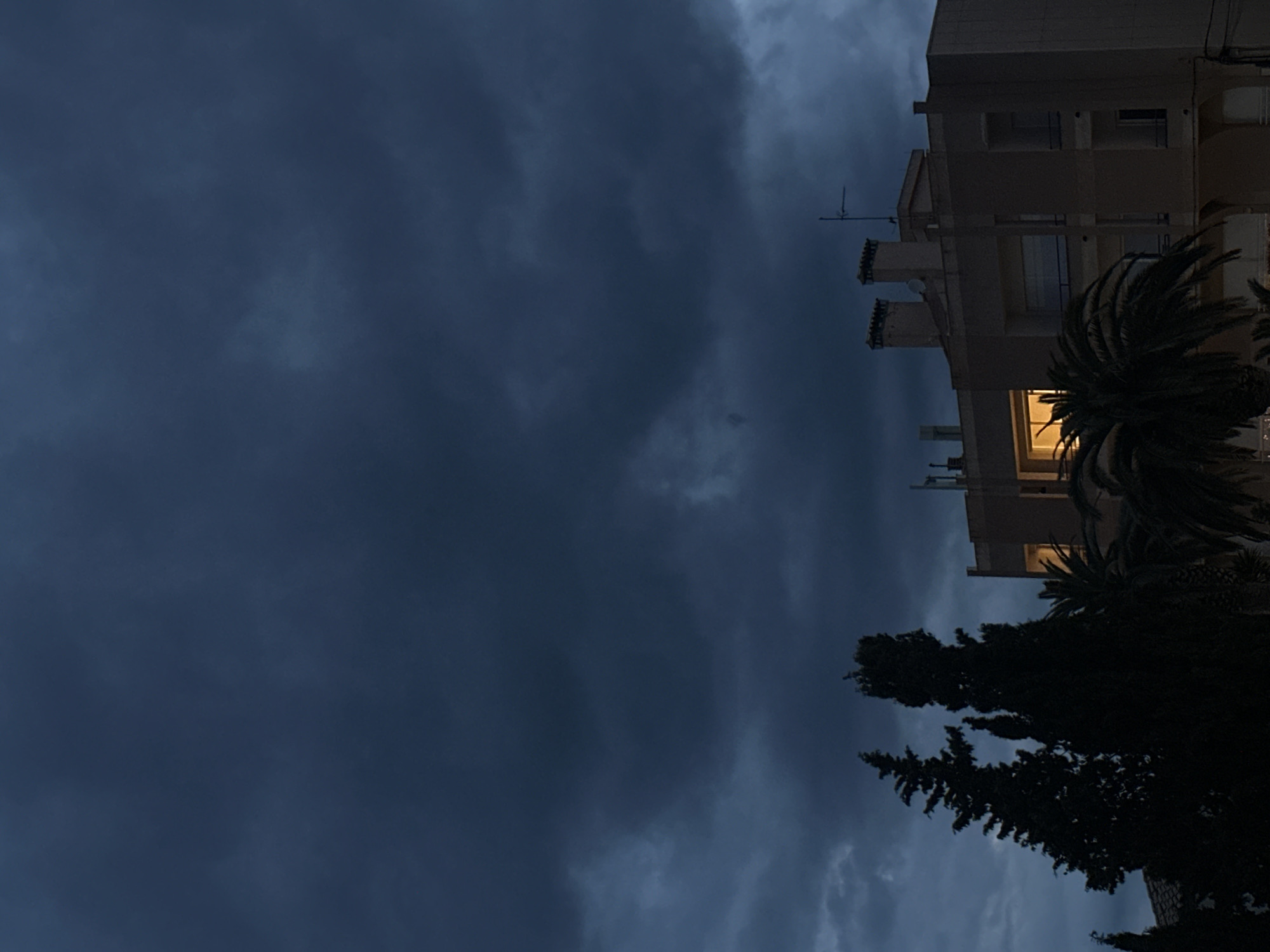 The image shows a building and a tree with a cloudy sky in the background. The building is tall and has multiple floors. A light is on in one of the apartments. The tree is dark and silhouetted against the sky. The sky is cloudy and dark. The image was likely taken in the city of Sant Feliu de Guíxols, Spain.