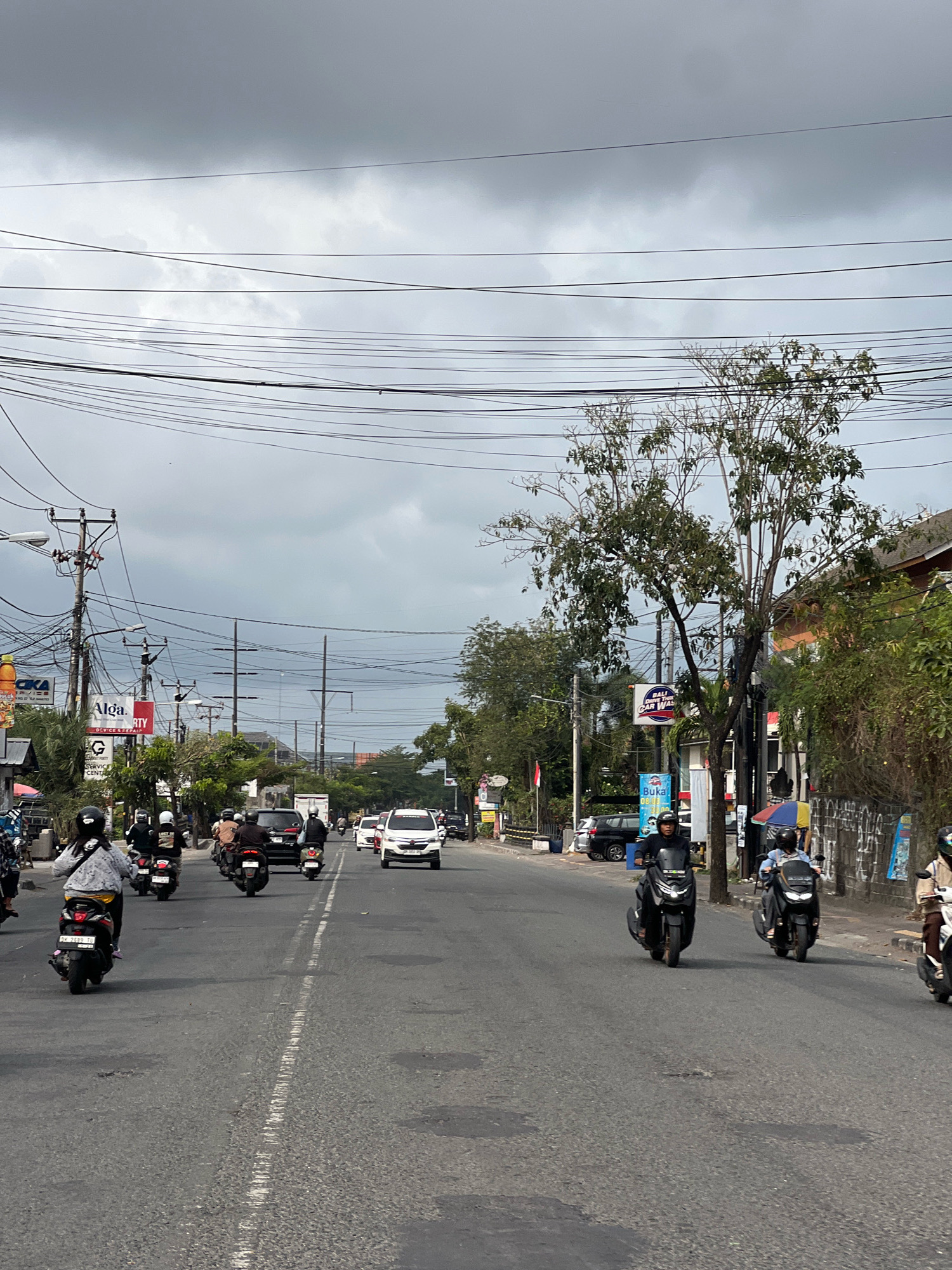 The image depicts a street scene in Denpasar, Indonesia, under a cloudy sky. The road is asphalt, with a visible white double yellow line marking the center. Several motorcycles are present, with riders wearing helmets. Some motorcycles are in motion, while others appear to be parked. Vehicles in the background include cars and other motorcycles. Buildings line the street, featuring storefronts and signage, including an "Alga. RTY DEVICE REPAIR" sign and a "BALI CAR WASH" sign. Power lines are prominently displayed overhead, strung across multiple poles. Trees and vegetation are visible along the sides of the road. The overall atmosphere is that of a typical urban street with ongoing traffic.