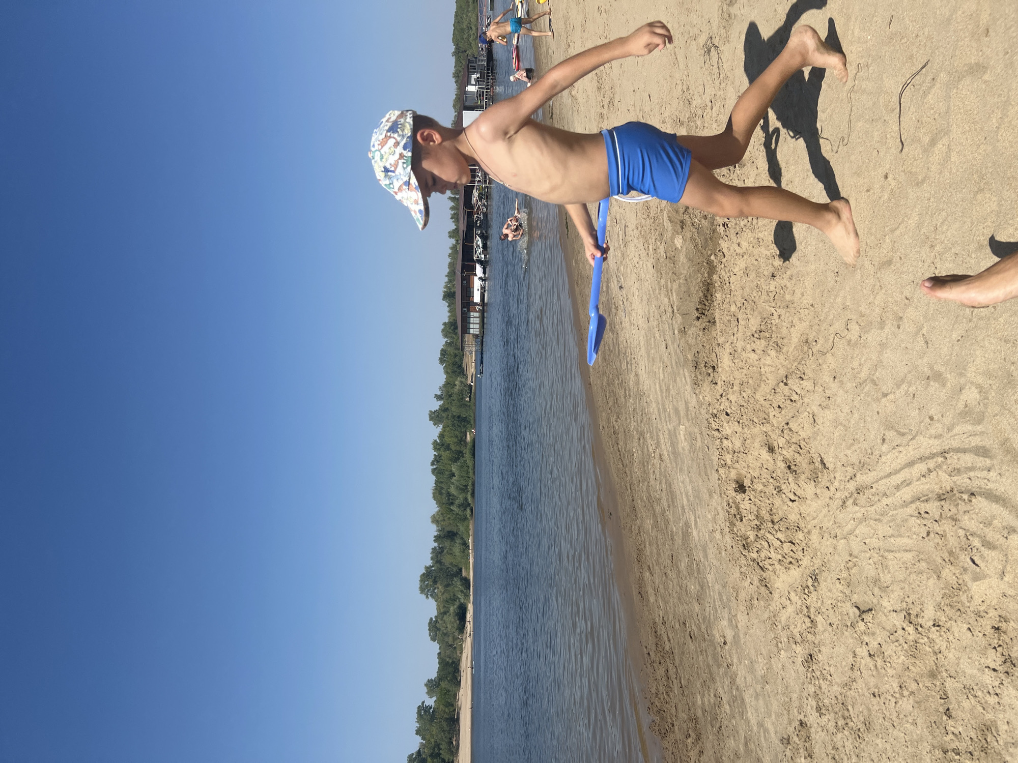The image shows a boy walking on a sandy beach next to a body of water, presumably the Dnieper River in Kilyakovka, Russia. The boy is wearing blue swim trunks and a patterned baseball cap. He is holding a blue shovel in his right hand. The sky is clear and blue. In the background, there are some buildings on the water and trees along the opposite bank. Other people can be seen in the water and on the beach. The sand is light brown and appears to be somewhat dry. The water is calm and reflects the blue sky.
