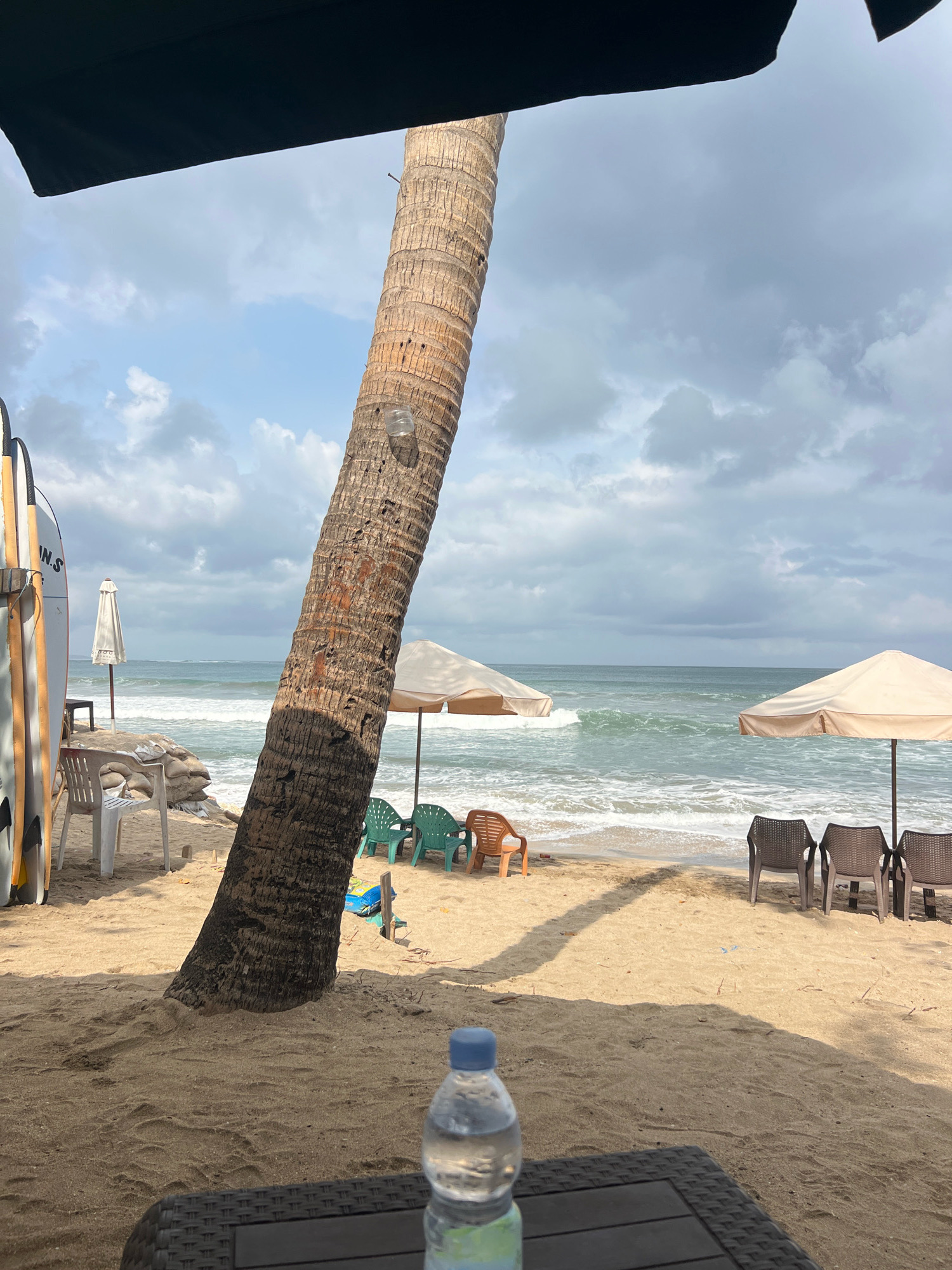 This is a photograph taken from a beach in Kuta, Indonesia, likely a resort area, as suggested by the presence of surfboards, beach chairs, and umbrellas. The foreground shows a sandy beach with a woven table and a partially visible plastic water bottle. A large palm tree trunk dominates the center of the image, casting a shadow on the sand.
In the midground, more of the beach is visible, with several beach chairs arranged under two large, light-colored umbrellas. Some of these chairs are occupied by people, though their features are not distinct. To the left, a stack of sandbags and a white plastic chair are visible. A white umbrella stands further back. Several surfboards are propped up against a wooden structure on the far left of the image.
The background features the ocean, with moderate waves breaking near the shore. The water is a bluish-green hue. The sky is cloudy, with patches of blue visible through the gray clouds. The overall atmosphere is one of a moderately busy beach, with elements of relaxation and water sports.