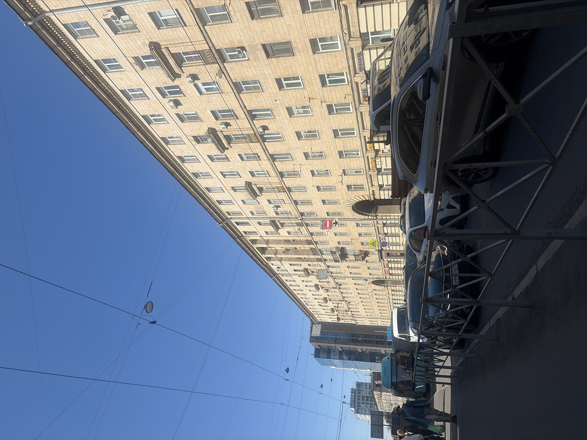The image shows a street in Saint Petersburg, Russia. A tall, light-colored building with many windows dominates the left side of the frame. Cars are parked along the right side of the street, partially obscured by a metal barrier. People are walking on the sidewalk, including a man with a backpack. Above, power lines stretch across a clear blue sky. Skyscrapers are visible in the background.