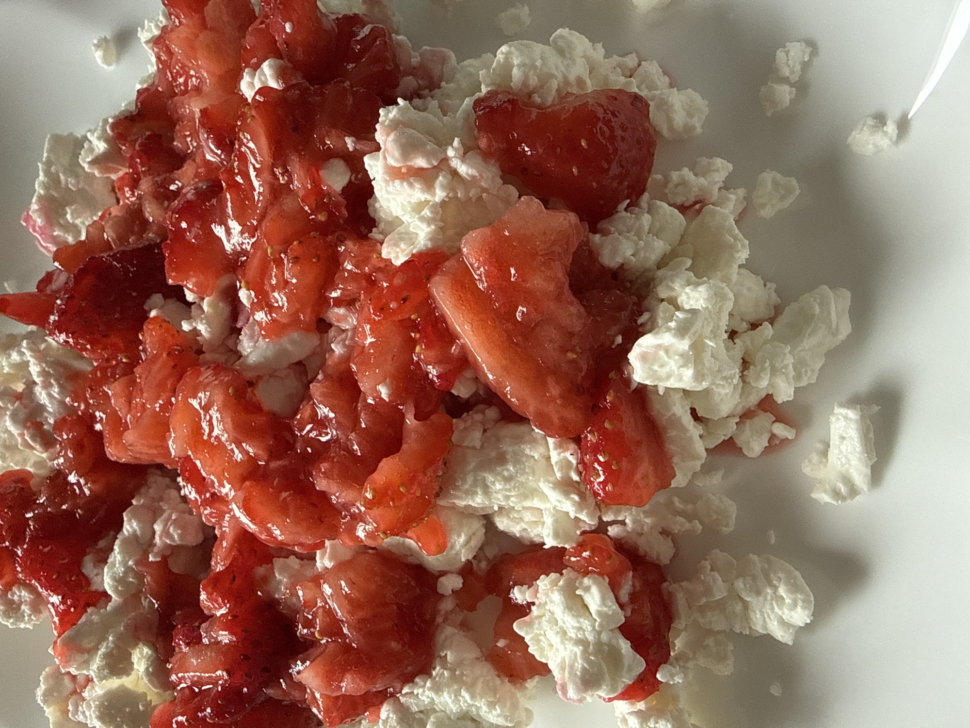 A close-up, overhead shot captures a mound of crumbled cottage cheese topped with sliced strawberries and strawberry sauce on a white plate. The strawberries appear to be fresh and juicy, some with visible seeds. The cottage cheese is fluffy and white, with some pinkish hues from the strawberry sauce. The plate is plain white and shows no markings. There are no people or other objects visible in the frame. The image is well-lit, with the lighting casting soft shadows on the food. This scene is not specific to Khimki, Russia, or any other geographical location.