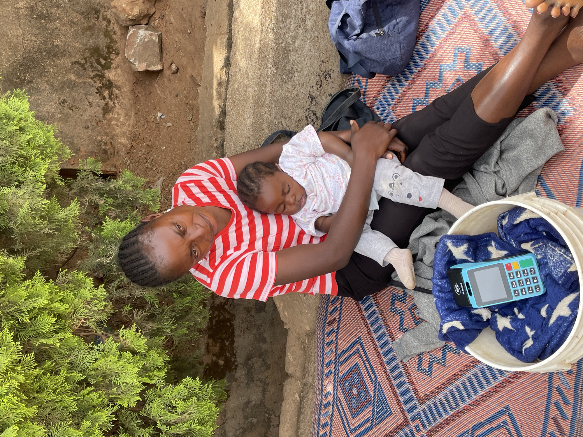 A daytime outdoor scene in Jos, Nigeria, features a woman cradling a sleeping baby. The woman, with dark skin and braided hair, wears a red and white striped t-shirt and dark pants. She is seated on a colorful patterned mat (red, blue, and white) spread across an earthy ground, which also shows rocks and concrete. She looks directly at the camera with a neutral expression.
The baby, nestled on her lap, appears to be asleep, wearing a light-colored patterned top, grey pants, and light-colored socks. To the woman's left, vibrant green shrubbery, resembling cypress, grows from the ground. Behind her on the mat, a blue backpack and a black bag are visible.
To the woman's right, a white bucket holds a blue blanket adorned with white star patterns. Inside the bucket, a blue and black Point of Sale (POS) machine is prominently displayed. The machine has a dark screen, a numeric keypad (0-9), and function buttons labeled F1 through F4. A "VERVE" logo is visible on the upper left corner of the POS device.
The bright, natural illumination suggests clear weather during the day. The image captures a moment of rest or waiting, with the woman attentively holding the sleeping child, and the presence of the POS machine hinting at potential business activities.