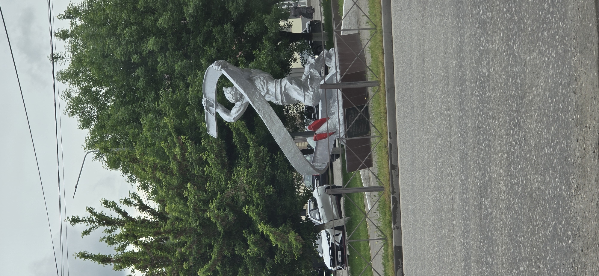 Here is a detailed description of the image:
**Location:** An outdoor setting, likely a city street in an unknown location.
**Subject:** The primary subject is a silver statue of a male figure appearing to operate a tool or machinery that forms a large curve. The statue is placed on a grey concrete base. There are two red objects on the base near the statue.
**Environment:** The statue is set against a backdrop of lush green trees, with the sky a light grey color. The area next to the statue is a grey paved street.
**Objects:** Besides the statue, the base it rests upon, the trees and street, there are some cars parked nearby. A decorative metal fence separates the statue from the road. Electrical wires are visible in the sky above the trees.