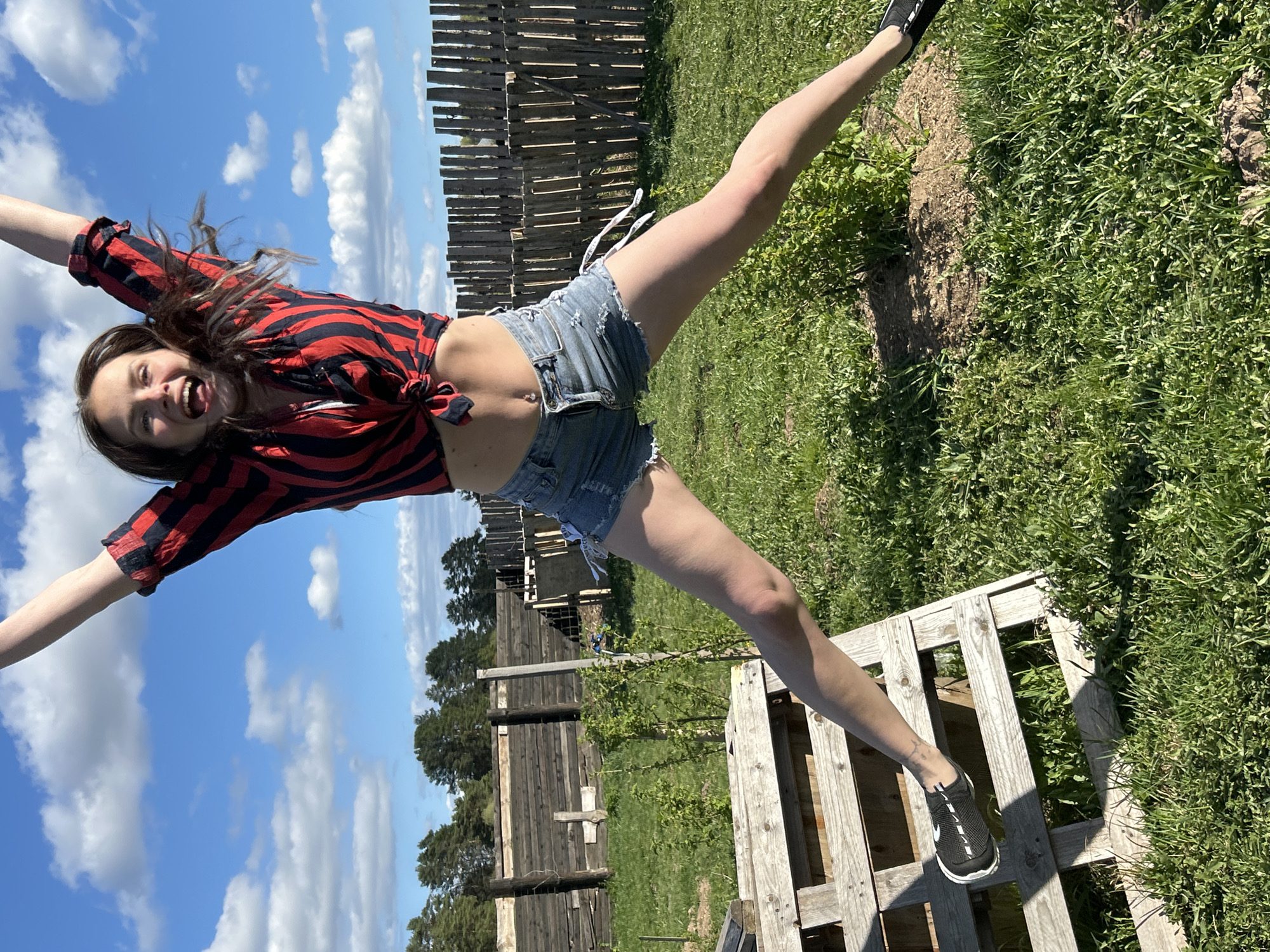 A young woman stands on a wooden pallet in a grassy area with a rustic wooden fence in the background. She is wearing a red and black striped shirt tied at the waist and denim shorts. Her arms are outstretched, and one leg is extended into the air. The sky is visible, with a mix of blue and white clouds. The scene appears to be outdoors in a rural setting, possibly in or near Rusalevka, Russia, given the location metadata.