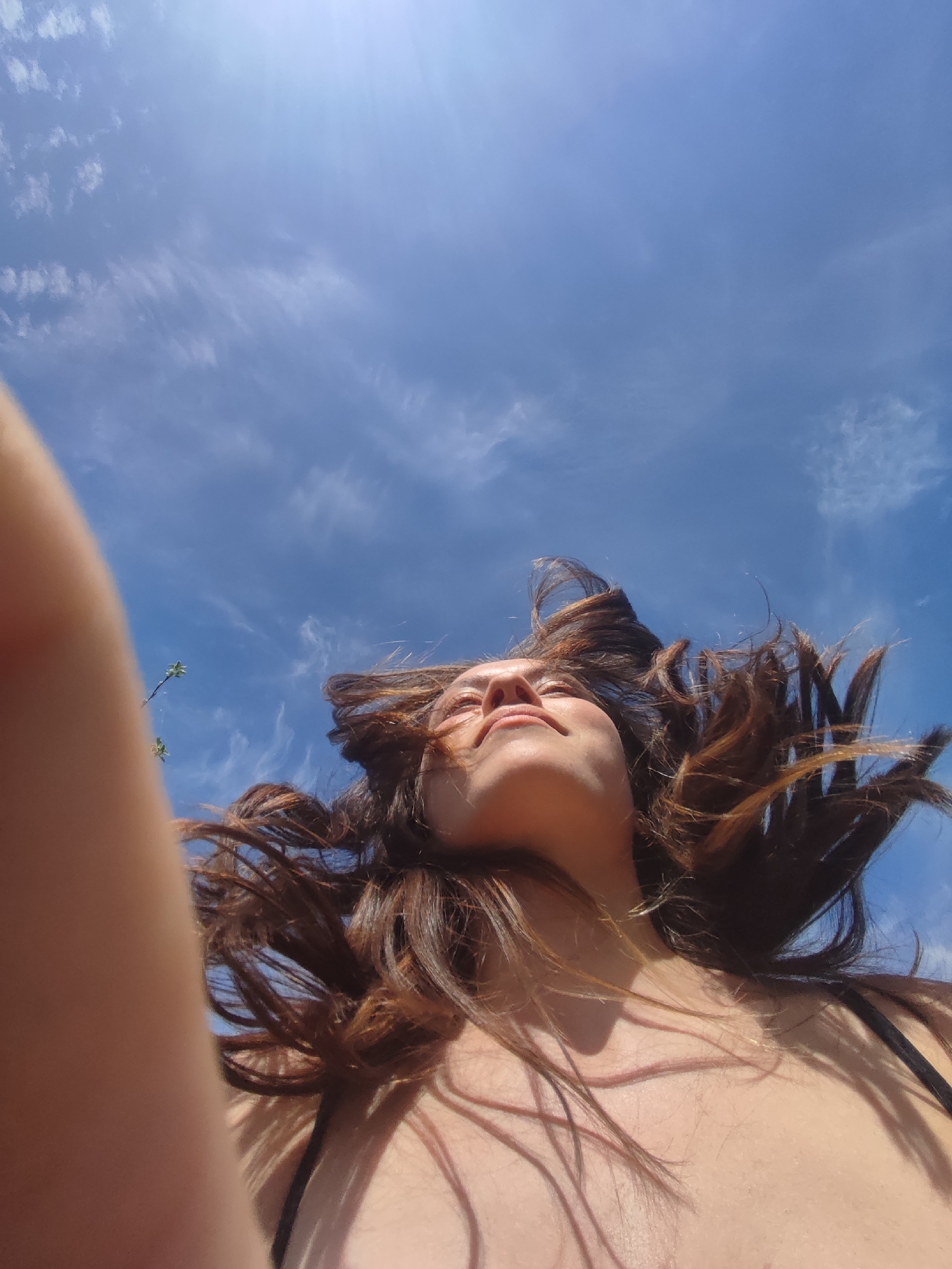 A low-angle, slightly off-center view captures a woman's face and torso against a bright blue sky with scattered white clouds. Her brown hair is windswept, obscuring parts of her face. The camera appears to be held by the subject, with a portion of the arm visible on the left edge of the frame. The sky is the dominant background, with slight traces of greenery visible at the very edge of the image. There is no indication of this being in Фоки, Russia.