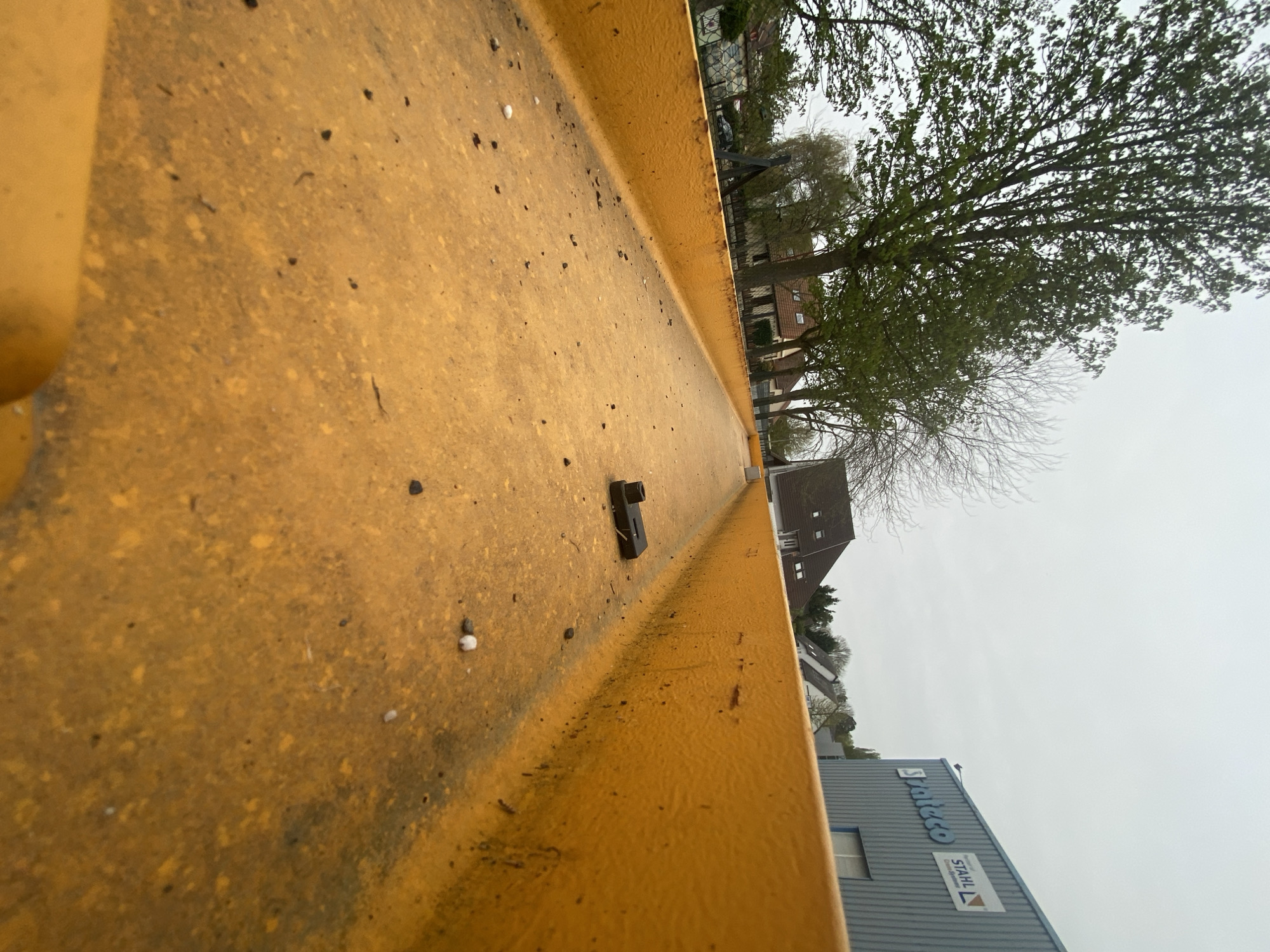 The image shows a close-up view of the interior of a yellow, potentially metal, container or structure. Debris, including small dark particles and a single white pellet, is scattered on the surface. A small, black, rectangular object with a raised feature is visible.
In the background, out of focus, is a street scene in Truchtersheim, France. Buildings, including houses with pitched roofs and a commercial building with "Stahlco" signage, are visible. Trees, some with sparse foliage, are present. The sky is overcast and grey.