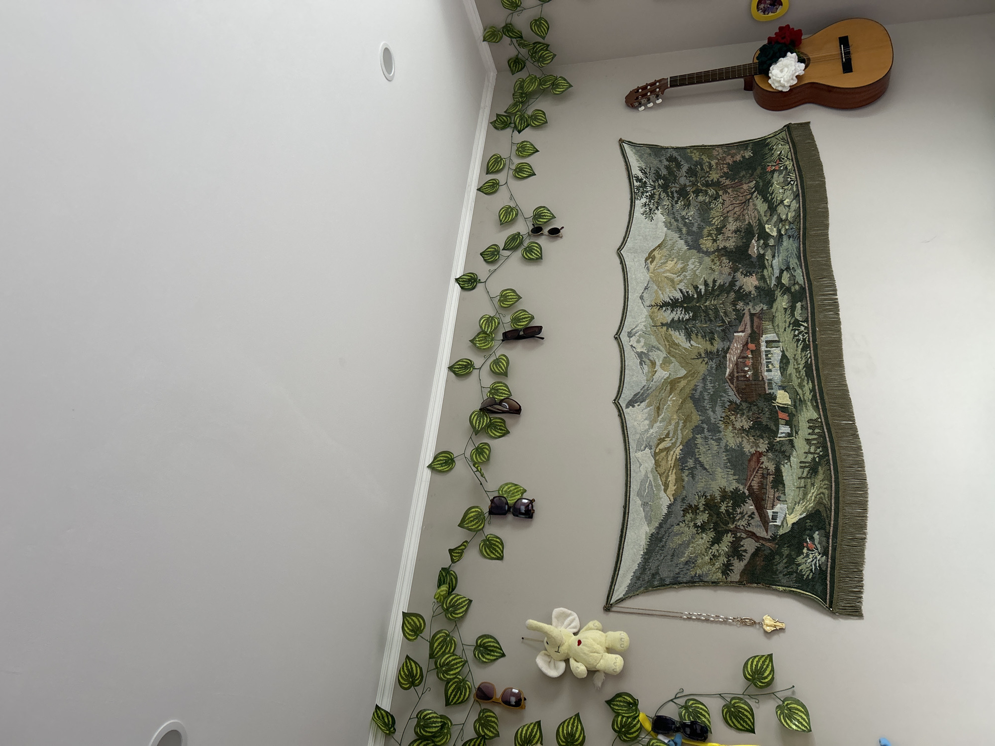 The image shows a wall decorated with various items. A tapestry depicting a mountain scene hangs on the wall. A guitar with floral decorations rests above the tapestry. An artificial vine with sunglasses scattered along it runs vertically along the wall. A stuffed elephant toy is placed near the bottom of the tapestry. The wall is painted a neutral color. The location in the city of Pazardzhik, Bulgaria is unknown.