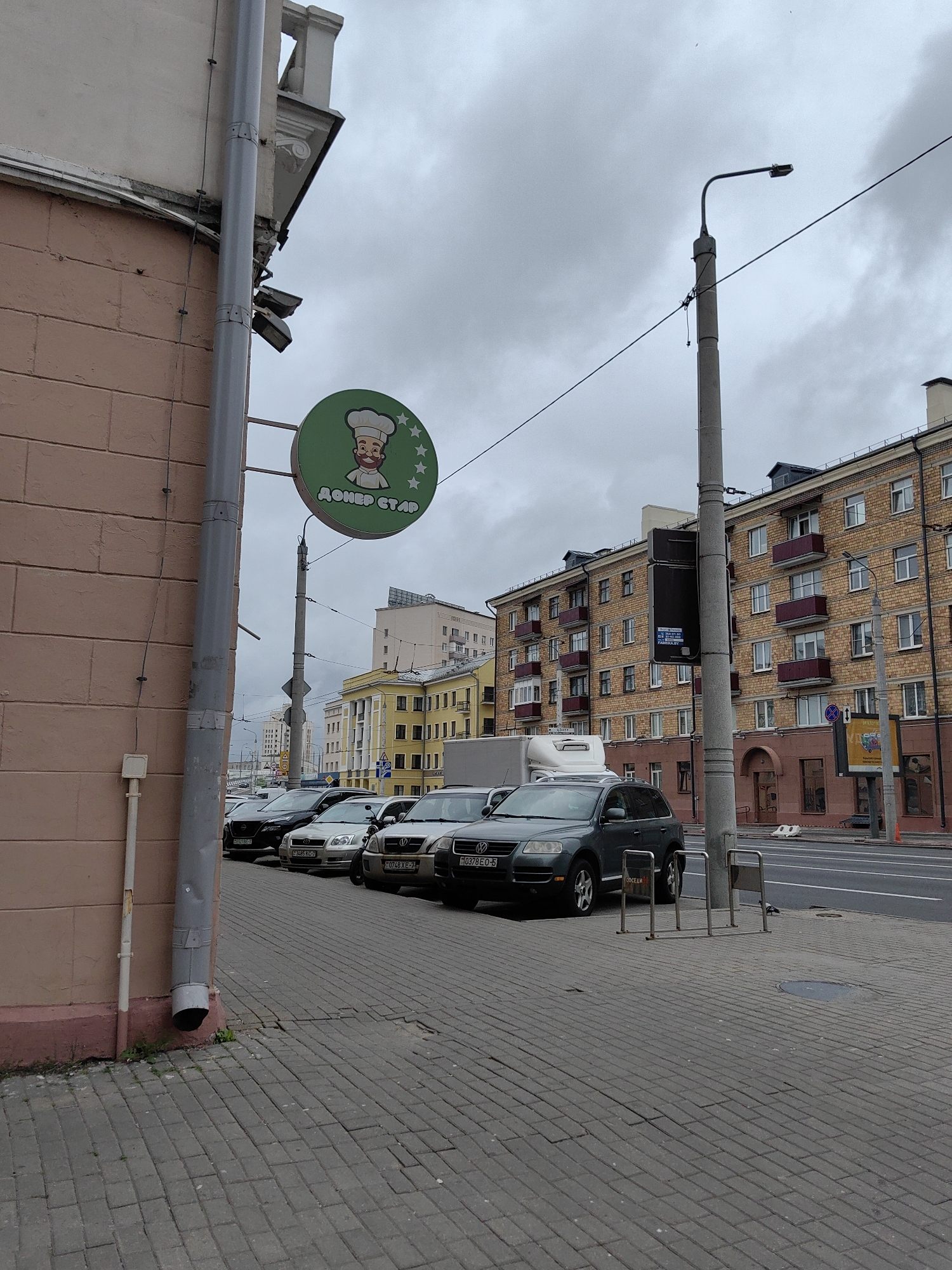 The image depicts a street scene in Minsk, Belarus, under an overcast sky. A light-pink brick building, possibly an apartment block or commercial property, occupies the left side of the frame, featuring a prominent grey downspout. A circular green sign with a cartoon chef and the text "ДОНЕР СТАР" (Doner Star) hangs from a pole.
In the foreground, a paved sidewalk with cobblestones leads to a street. Several cars are parked along the street, including a dark-colored SUV on the right and various sedans and SUVs in the background. A traffic light pole with a black traffic signal box stands near the center of the image.
Further in the background, a multi-story brick building with a red-brick facade and balconies is visible, along with other urban structures. The overall atmosphere is that of a typical city street on a cloudy day, with no people clearly visible.