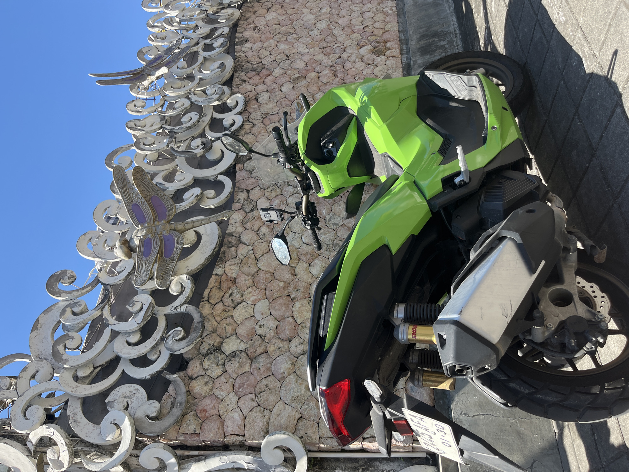 A vibrant green scooter is parked on a cobblestone pathway in front of a textured stone wall topped with an ornate, white, wave-like metal railing. The scooter's rear is angled towards the viewer, showcasing its red taillight, silver exhaust pipe, and suspension components. A license plate with the inscription "01-30" is visible. The wall features decorative dragonflies made of purple and gold metal. The scene is brightly lit by sunlight, casting shadows on the ground. The location is identified as Sukawati, Indonesia.