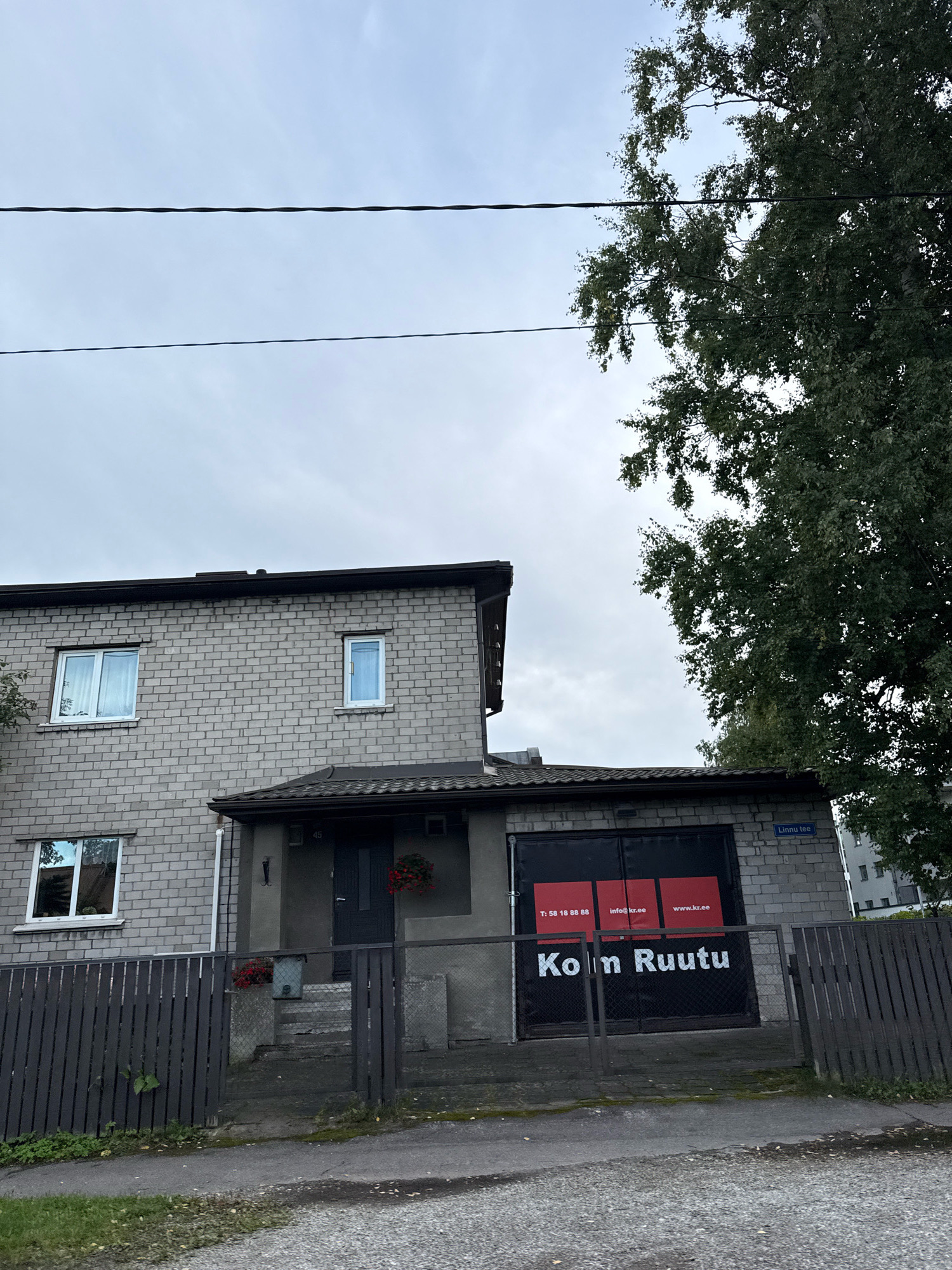 The image shows a two-story building with a grey brick facade and a dark-tiled roof. There is a garage door on the ground floor with a large black banner that reads "Kolm Ruutu" in white lettering. Below this, there are three red squares with white text: "T: 58 18 88 88", "info@kr.ee", and "www.kr.ee". A street sign above the garage door reads "Linnumäe tee". A dark wooden fence runs along the front of the property, with a gravel driveway leading to the garage. The environment appears to be a residential or commercial area in Tallinn, Estonia. A large tree with green leaves is visible to the right of the building, and overhead power lines are present in the upper part of the image. The sky is overcast.