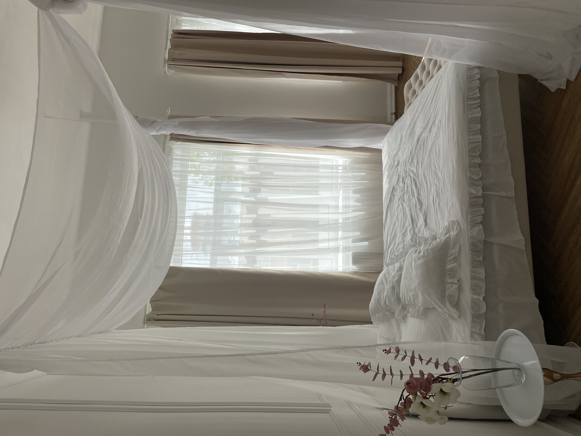 The image shows a bedroom interior. A white canopy bed is the main subject, with white bedding and pillows. A vase with pink and white flowers lies on a white surface near the bed. In the background, there is a window covered with sheer white curtains and beige blinds. The floor is made of wood. The image does not provide any information about the location being Kazan, Russia, or the presence of any people, actions, or interactions.