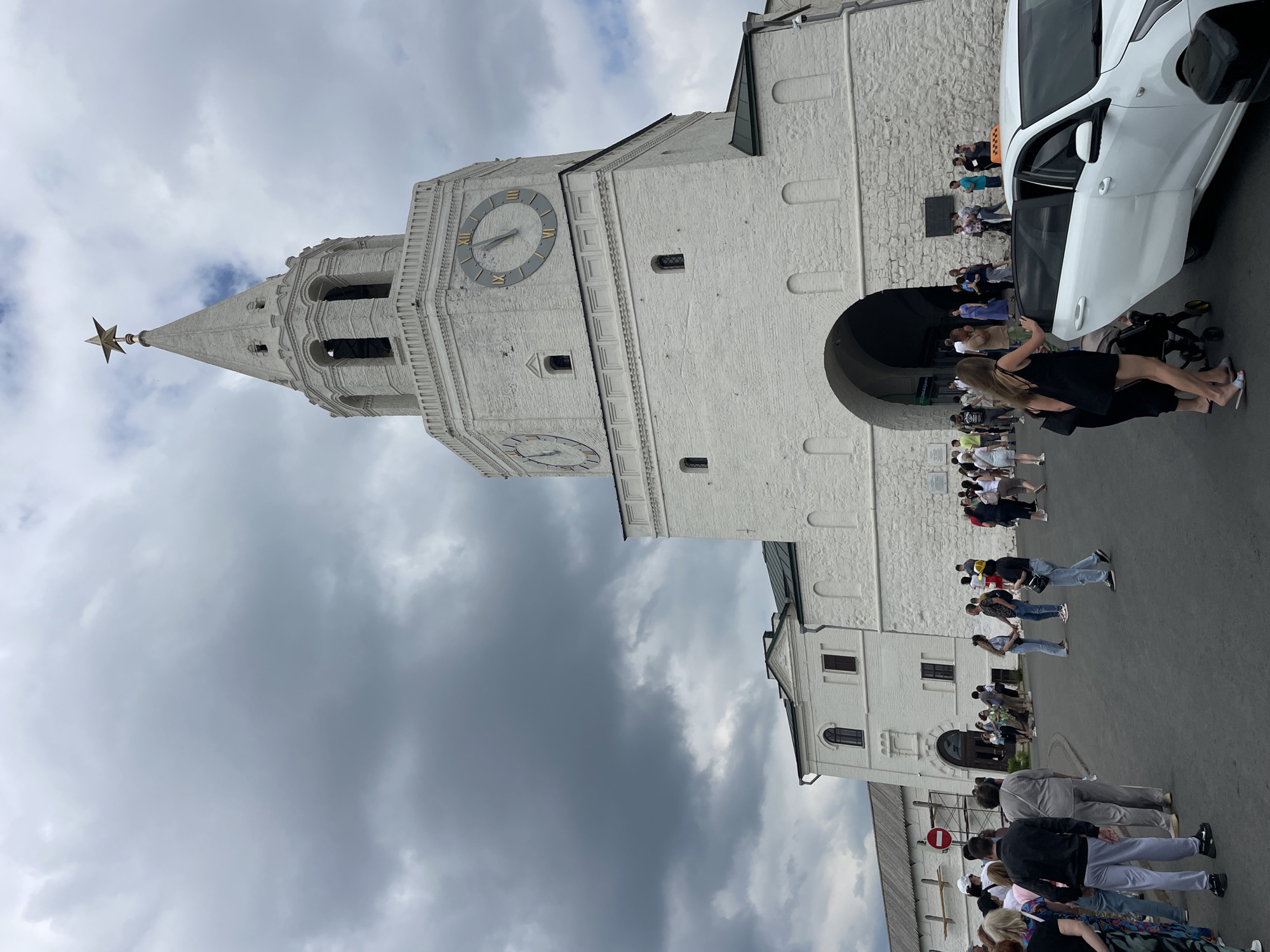 In Kazan, Russia, a wide shot captures a bustling scene at the Kazan Kremlin, focusing on the iconic Spasskaya Tower. The imposing white brick tower, topped with a golden star, dominates the frame. Two large clock faces adorn its sides, with Roman numerals indicating the time. Dark, dramatic clouds gather in the sky, creating a moody atmosphere. A crowd of tourists and locals mills about in front of the tower's grand arched entrance, some in the process of entering, others pausing to admire the architecture. To the right, a white car, possibly a taxi, is parked with its doors open, suggesting arrivals or departures. The foreground shows people dressed in casual summer attire, some carrying bags or pushing strollers. The scene conveys a sense of activity and historical grandeur.