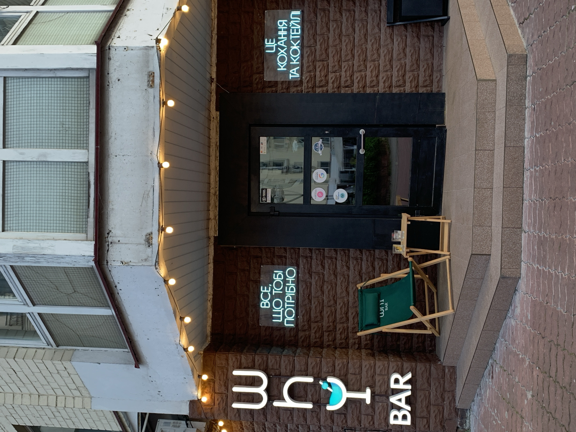 Here is a description of the image:
The image shows the storefront of a bar located in Kyiv, Ukraine. The name of the bar is "Wh111 Bar," as indicated by the illuminated sign at the bottom of the image. Above the entrance, there are two neon signs in Ukrainian: one says "Це кохання та коктейлі" (This is love and cocktails) and the other says "Все, що тобі потрібно" (Everything you need). A black door leads into the bar. Near the entrance are a small table and a green folding chair with the bar's name on it. The exterior walls are brown with a brick-like texture. The sidewalk in front of the bar is paved with reddish-brown bricks. A string of lights hangs along the awning of the building to the left of the bar. The building appears to be multi-story.