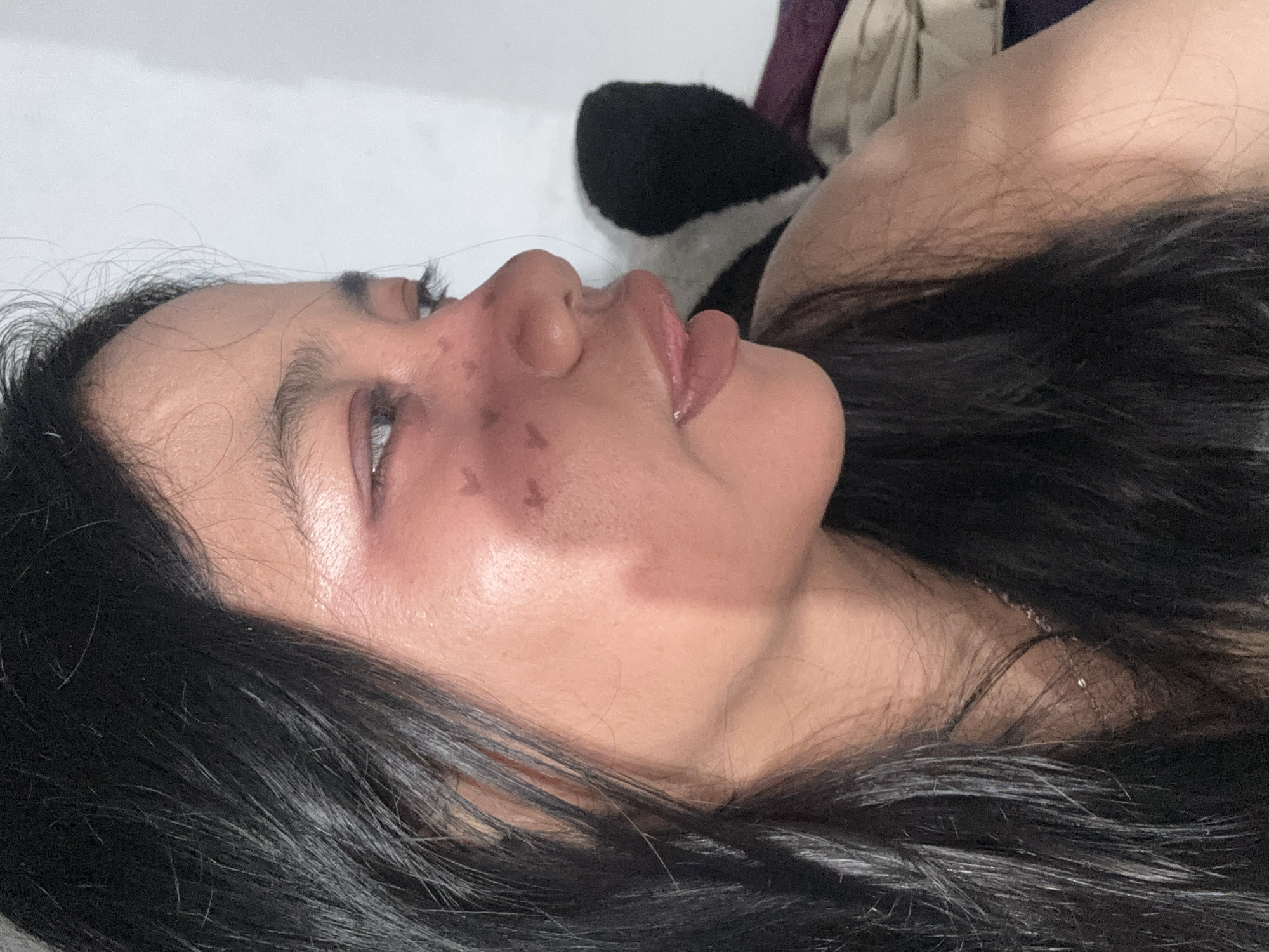A close-up profile shot captures a young woman with dark hair and fair skin, her head turned to the right. Her left eye is visible, looking down and to the right, with apparent makeup on her eyelid. Several small, dark, butterfly-shaped markings are visible on her cheek and nose. Her lips are slightly parted, revealing a hint of her tongue. The background is a plain, white wall, with a dark, fuzzy object partially visible behind her right shoulder. The overall impression is of a self-portrait taken indoors. The image does not contain enough information to determine its specific location within North Jakarta, Indonesia.
