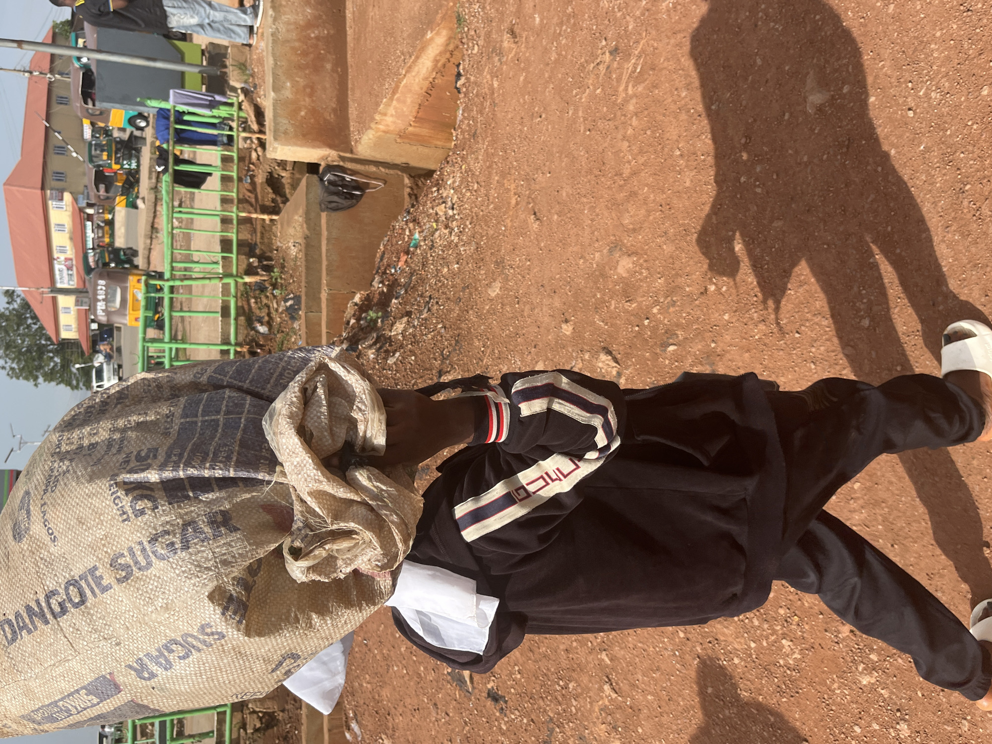 A person, likely a child based on the size of the bag and the overall proportions, is walking away from the camera, carrying a very large sack of sugar on their head. The sack has "DANGOTE SUGAR" printed on it. The person is wearing a black hooded jacket with striped sleeves and dark pants, and white sandals. They are walking on a dusty, reddish-brown dirt path. In the background, there is a busy street with several auto-rickshaws, a building with a red roof, and a green fence. There is also a pile of debris to the right of the path. The lighting suggests it is daytime. The specific location is identified as Gura Top, Nigeria.