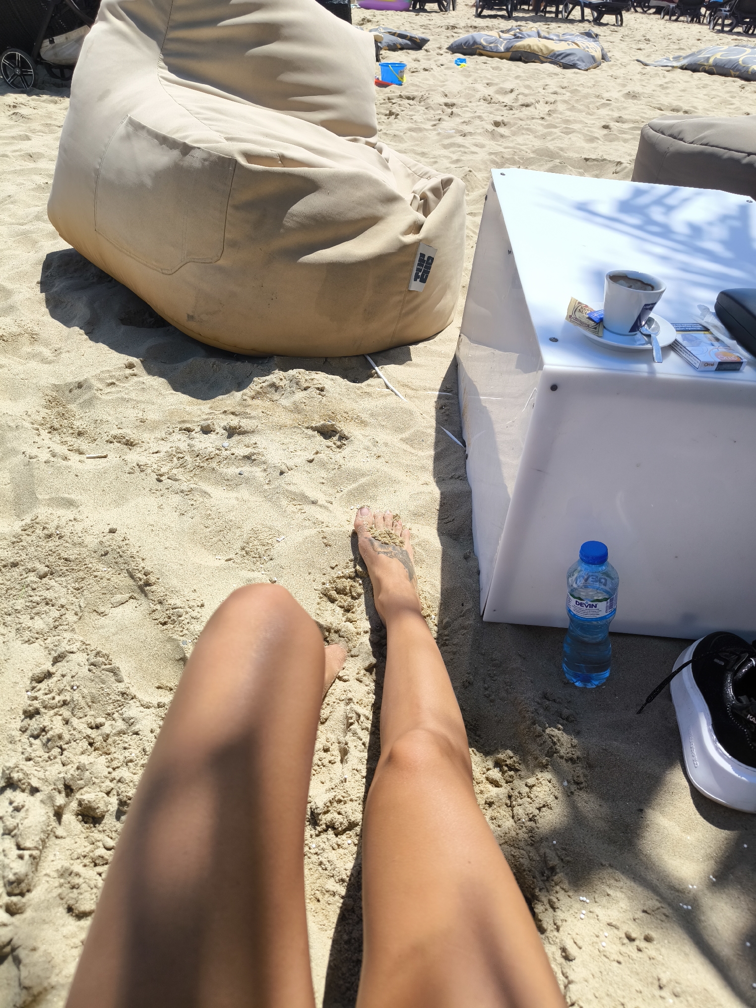 The image depicts a sunny day on a sandy beach, likely in Nesebar, Bulgaria, given the presence of beach chairs and umbrellas often seen in tourist areas. In the foreground, a person's legs and feet are visible, resting on the sand. The feet have intricate dark tattoos. To the right, a modern white table holds a cup of coffee, a packet of sugar, a spoon, and a pack of cigarettes, suggesting a moment of relaxation. A blue bottle of Devin mineral water stands beside the table, and a black and white sneaker is also present. In the background, large beige beanbag chairs are scattered across the sand, characteristic of a beach lounge area. Further back, rows of empty sun loungers and more beanbag chairs are visible, with other beachgoers and their belongings faintly discernible. The overall scene conveys a sense of leisure and enjoyment in a coastal resort setting.