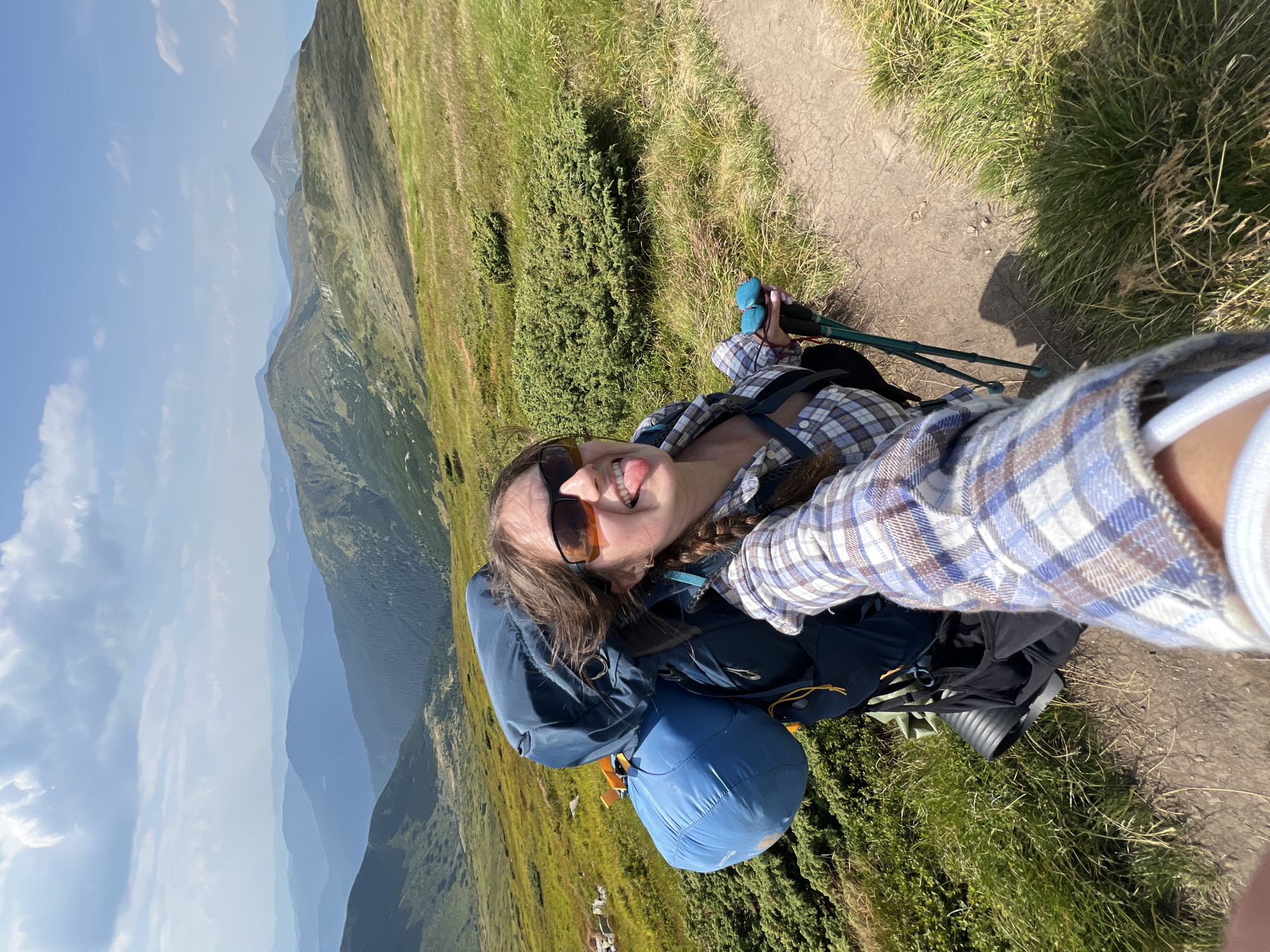 A young woman is taking a selfie while hiking in the Carpathian Mountains near Luhy, Ukraine. She is wearing a plaid shirt and sunglasses, and her tongue is sticking out. She has a large backpack on and is holding hiking poles. The background shows rolling green hills and mountains under a bright blue sky with white clouds. The trail she is on is dirt and rocky, with grass and bushes on either side.
