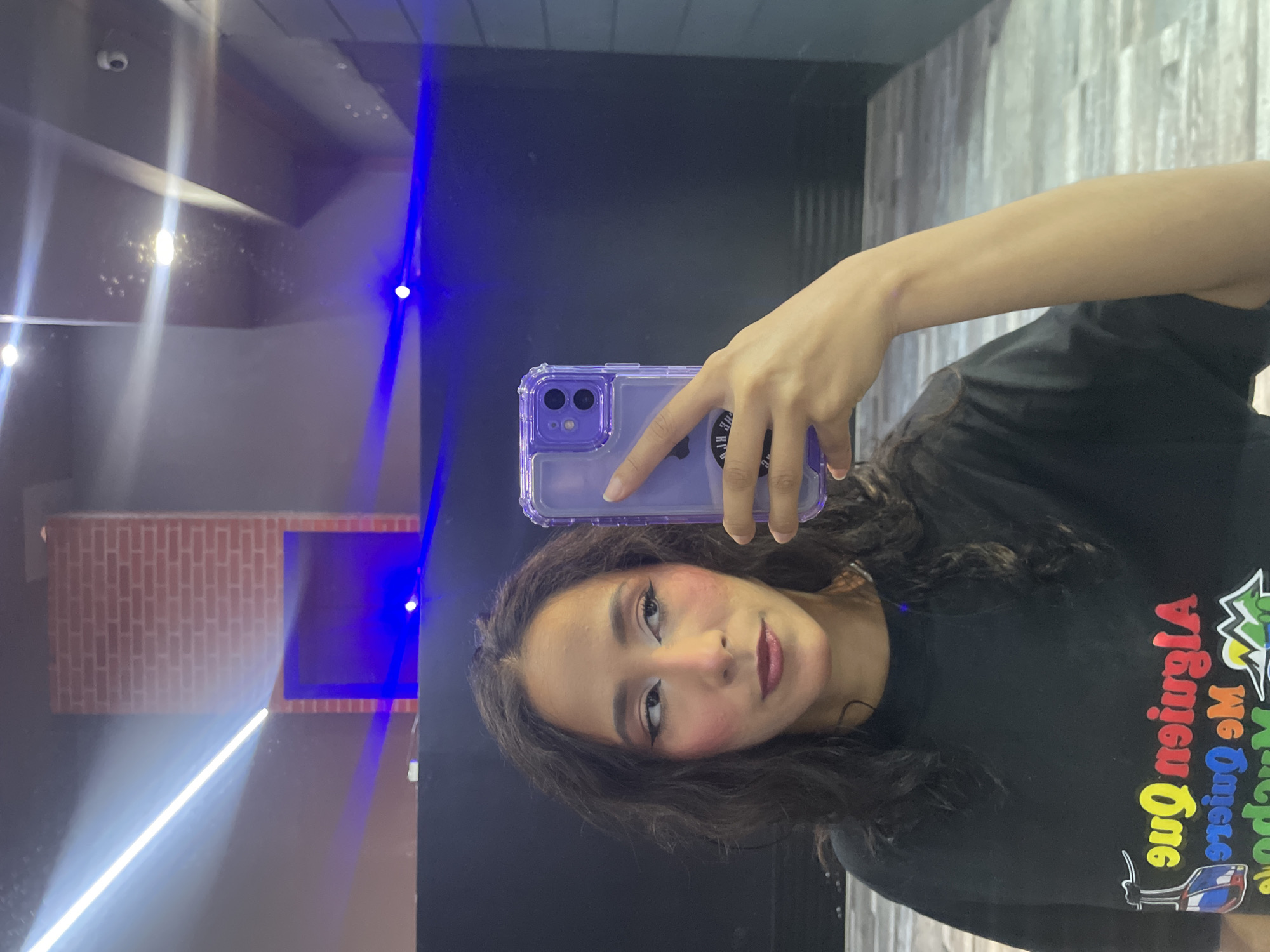 A woman with dark, wavy hair is taking a selfie in a room with a black wall and blue lighting. She is wearing a black t-shirt with colorful text and graphics on the front. The text on her shirt appears to be in Spanish. Her makeup includes winged eyeliner and blush. She is holding up a purple phone case. In the background, there is a faux brick fireplace and ceiling lights. The floor has a wood-like pattern. The environment appears to be an indoor space, possibly a studio or a club, given the lighting and reflective surfaces. The image was taken in Pampatar, Venezuela.