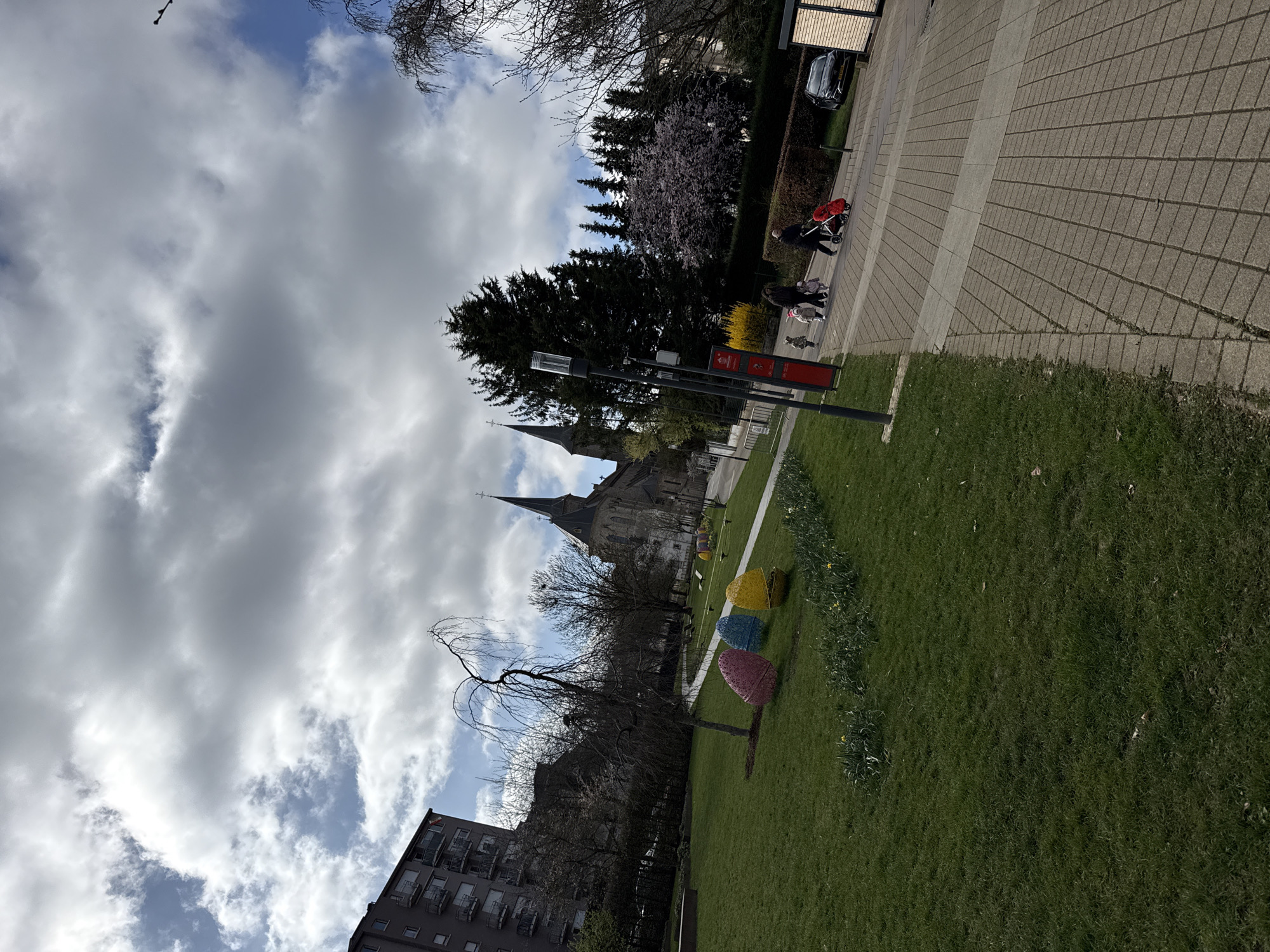 The image depicts an outdoor urban scene in the city of Unknown, Unknown, under a partly cloudy sky with large grey clouds and visible patches of blue. The foreground features a vibrant green grassy area, sloping gently upwards from left to right. Along the edge of the grass, a line of yellow daffodils is in bloom. Three large, decorative, egg-shaped objects, colored pink, blue, and yellow respectively, are prominently displayed on the grass. A slender, bare-branched tree with drooping limbs stands near these decorations.
To the right, a grey paved pedestrian walkway, constructed from rectangular pavers, runs parallel to the grassy area. A light grey stripe bisects the walkway lengthwise. On the far right of the walkway, a tall dark grey light pole with multiple fixtures stands adjacent to a red information kiosk or signpost.
In the midground, a group of five individuals is walking along the paved path from right to left. One adult, dressed in dark clothing, is pushing a red stroller or pram. Three small children, appearing to be toddlers or preschoolers, walk in front of the stroller. Another adult in dark attire walks alongside them, accompanied by a small, light-colored dog on a leash. Behind these individuals, a dark-colored car is parked on what appears to be a street or driveway, bordered by a dark green hedge and evergreen trees.
Further into the background, a large, historic stone church with two prominent spires dominates the skyline. To the left of the church, a multi-story residential building with grey facades and balconies is visible. A small flag, possibly red and yellow, hangs from one of its balconies. The scene also includes several trees, some with bare branches indicative of early spring, while one tree exhibits pink or purple blossoms.