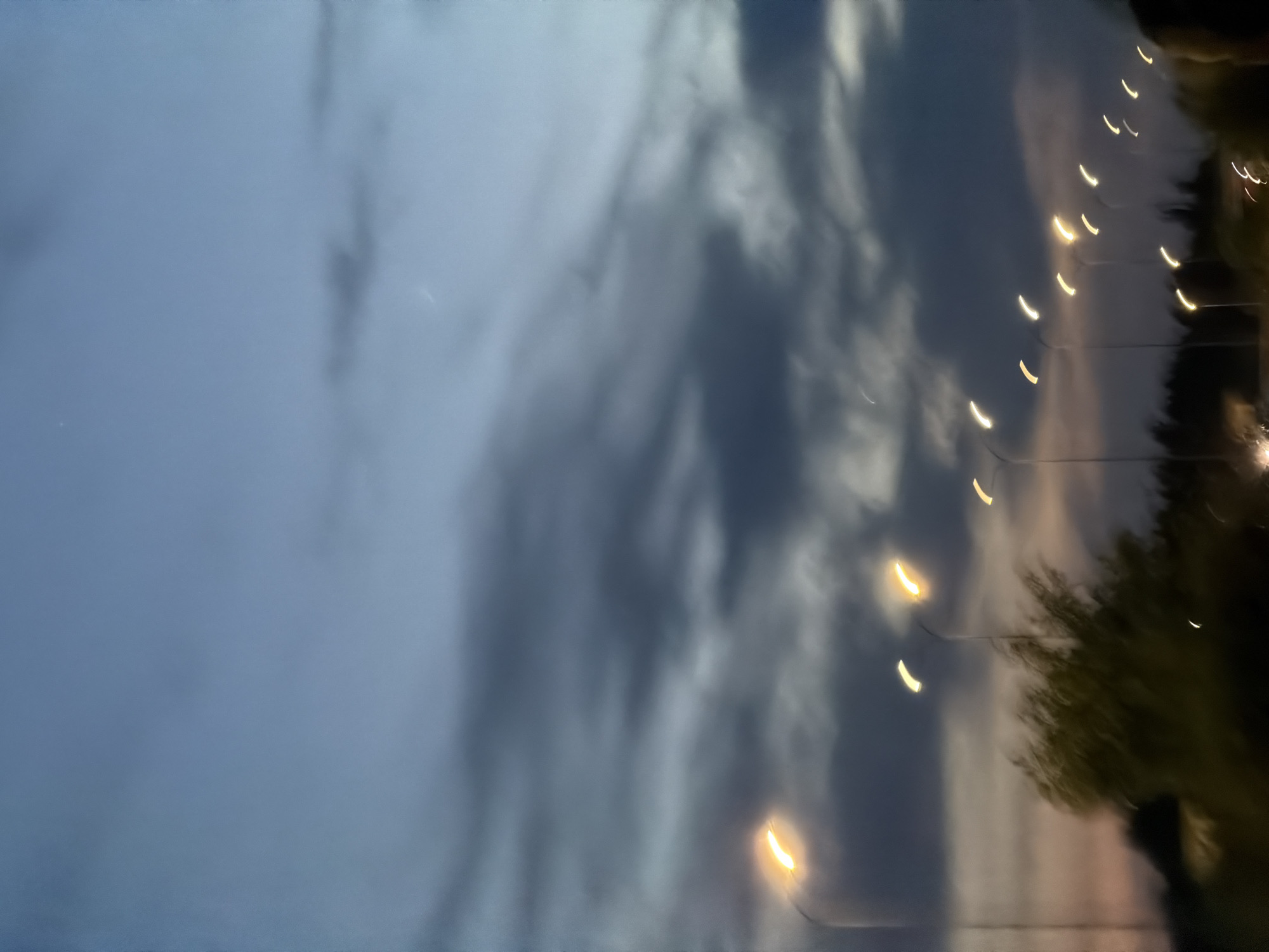 This is a vertically oriented, motion-blurred image taken at dusk or dawn in Kmytiv, Ukraine. A line of streetlights stretches across the mid-ground from left to right, each emitting a glowing orange-yellow light. The sky above is a gradient of blues and grays, with dark, wispy clouds obscuring parts of the horizon. Below the streetlights, silhouetted trees and foliage are visible, suggesting a roadside or park setting. The overall impression is one of movement and a transition from day to night. There are no discernible people or specific actions depicted, only the static elements of the environment blurred by motion.