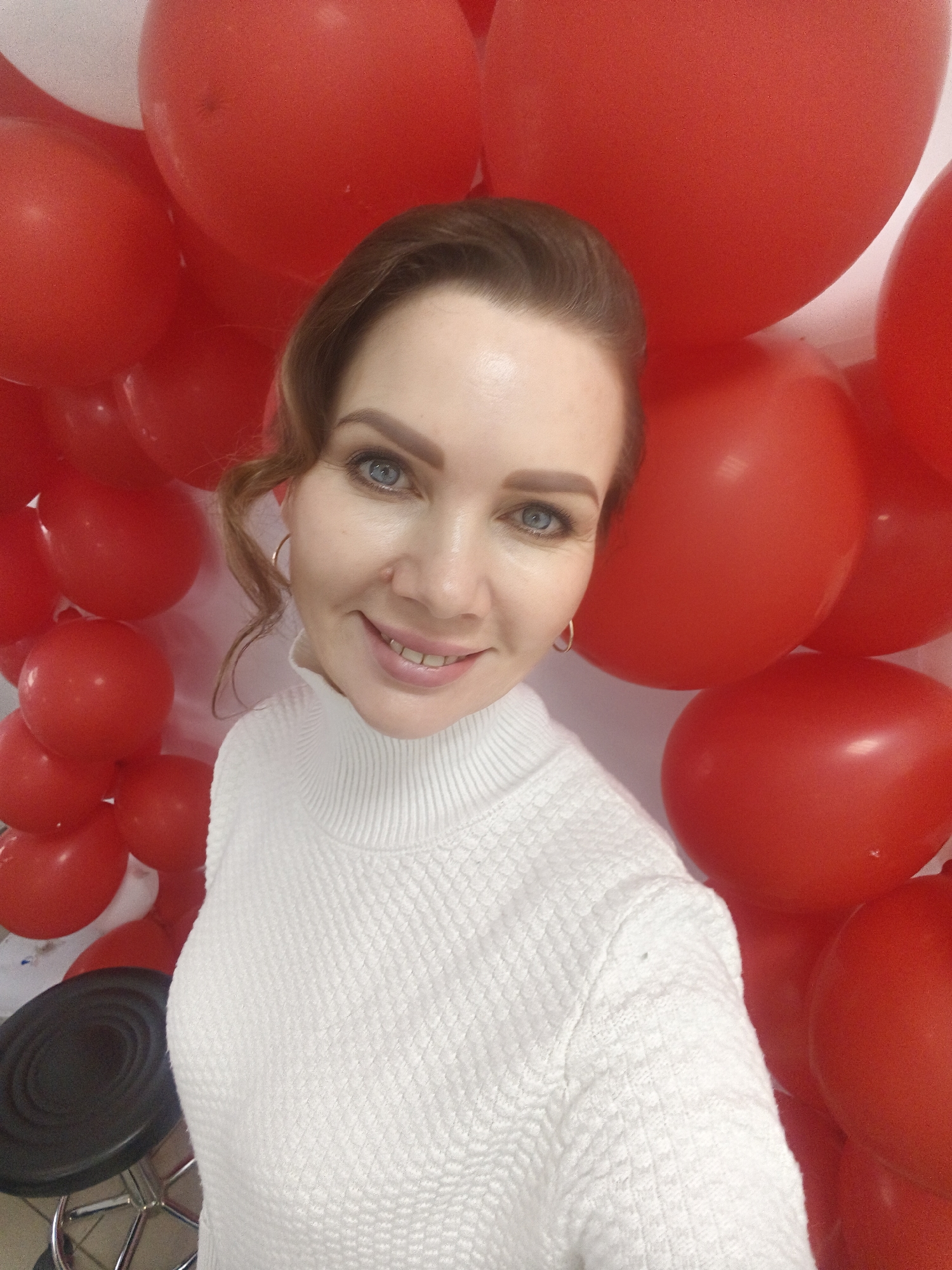 A woman is posing for a selfie. She has light skin, blue eyes, and her hair is pulled back in a loose style. She is wearing a white turtleneck sweater and gold hoop earrings. The background is filled with red balloons. Part of a black chair is visible in the lower left corner of the image. The location is Unknown, Unknown.