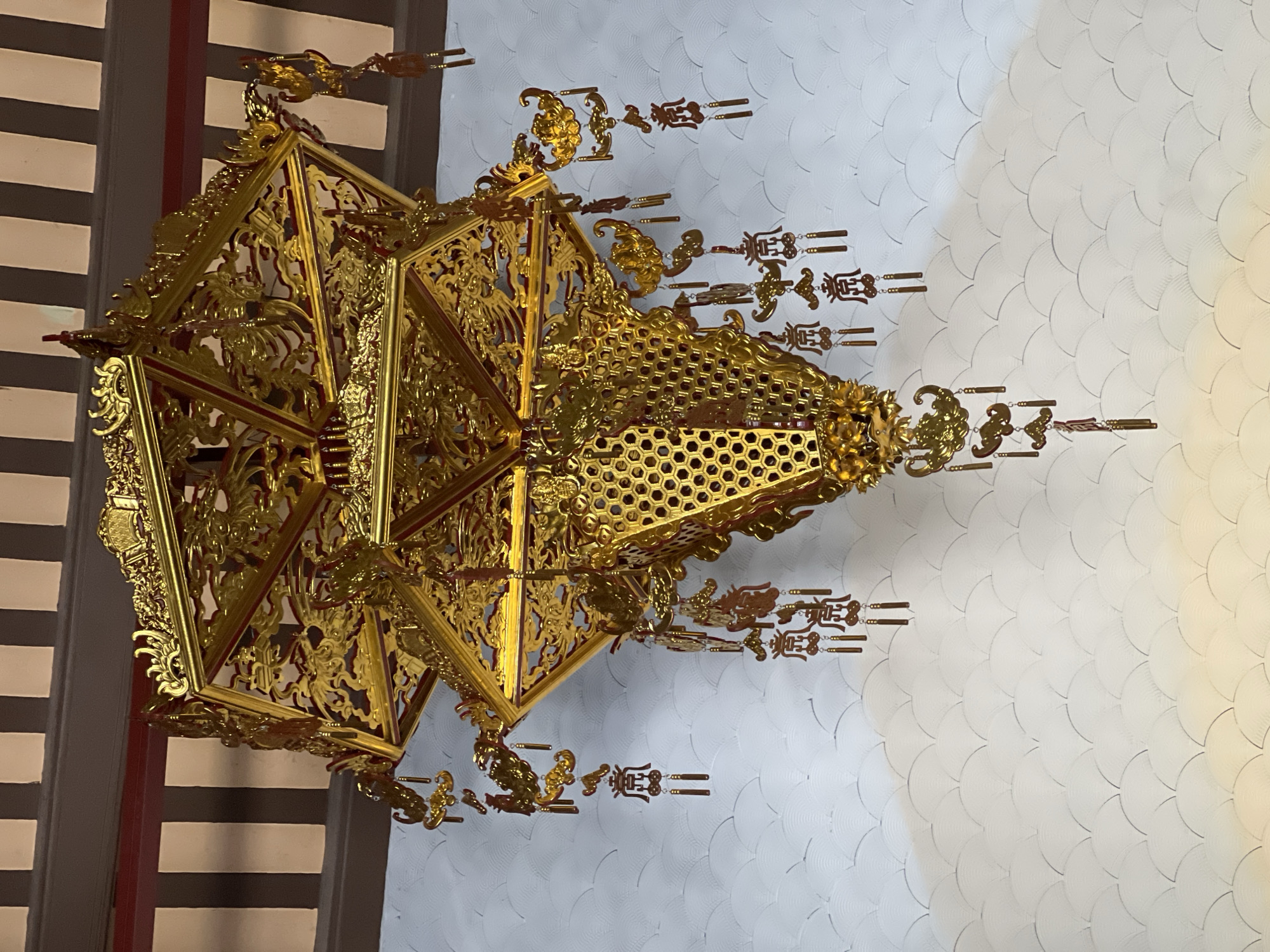 A traditional, ornate, multi-tiered golden lantern hangs from a wooden ceiling. The lantern is intricately decorated with cut-out patterns and small dangling ornaments, some of which appear to be calligraphy. The background wall features a repeating, scalloped pattern in a light color. The image appears to have been taken in the city of Đà Nẵng, Vietnam, likely in a temple or a similarly decorated cultural site.