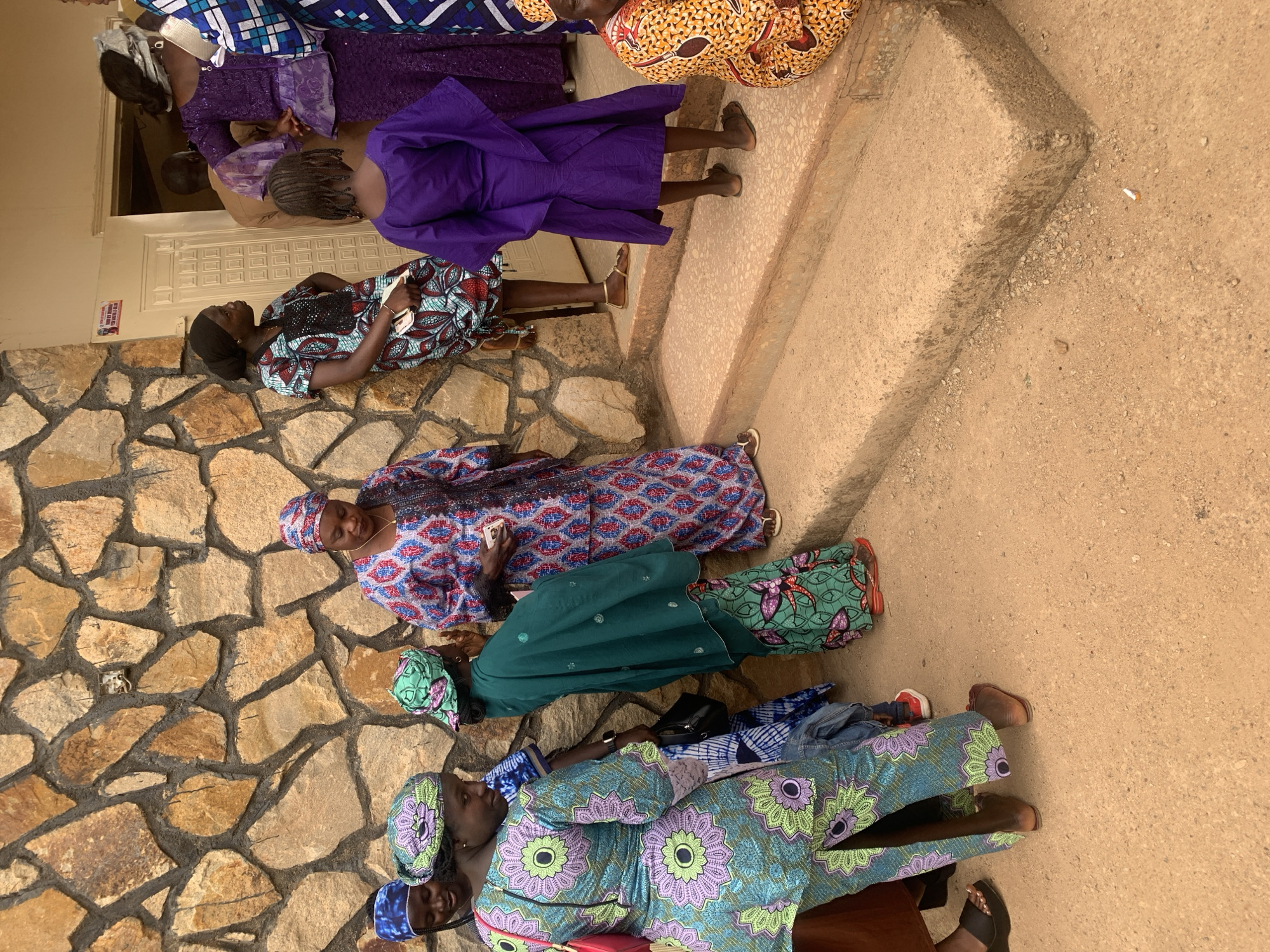 A group of women wearing colorful, patterned dresses and headwraps are gathered outside a stone building, possibly waiting for an event. The building is located in Jos, Nigeria. Some women are seated while others are standing near the entrance. The women's clothing is vibrant and diverse, suggesting a cultural gathering. The setting appears to be a sunny day, as indicated by the lighting and shadows.