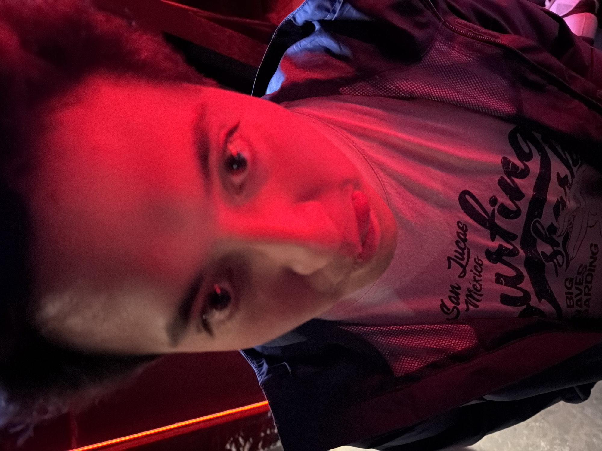 A person's face is illuminated by a red light. The person is wearing a jacket over a t-shirt. The t-shirt has an image of a surfer, with text that says "San Lucas Mexico Surfing Big Waves Boarding." The image does not contain sufficient information to determine the location in San Andrés Cholula, Mexico.