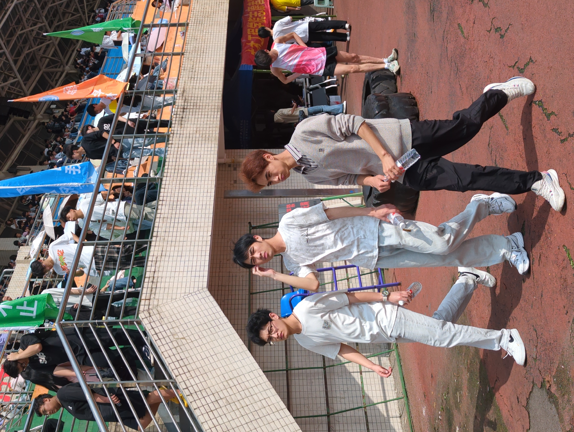 The image depicts an outdoor scene with a crowd of people in a stadium setting, possibly in Jiangjin, China. There are three young men in the foreground, each holding a plastic water bottle. One is wearing a grey sweater and black pants, while the other two are wearing white t-shirts and light-colored jeans. In the background, a stadium seating area is visible with spectators, flags, and banners in varying colors like orange, blue, and green. A building is seen behind them, and the ground appears to be a red pavement with patches of green. Some individuals can be seen walking on the ground while others are standing and engaging with each other.